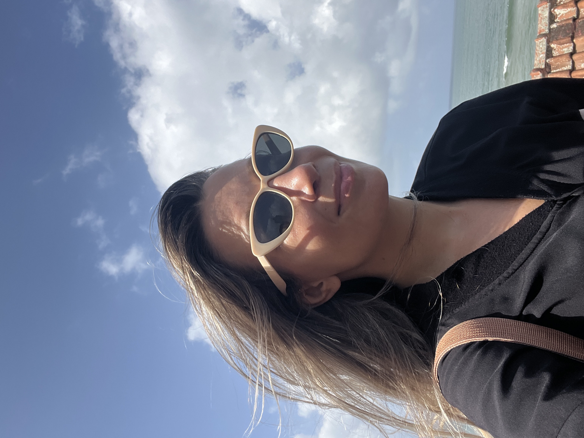 A woman wearing sunglasses and a black jacket stands with the ocean in the background. She has long, light brown hair that is being blown by the wind. The sky is blue with some white clouds. The woman is looking off to the right side of the frame. There is a terracotta tiled roof in the foreground. The scene appears to be in Walgama, Sri Lanka.
