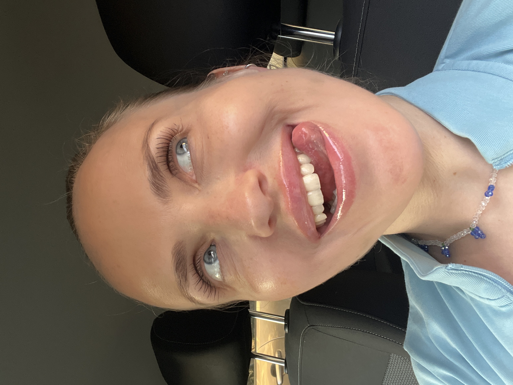 A young woman with blue eyes, fair skin, and blonde hair pulled back is in the driver's seat of a car. She is looking up and to her right with her mouth open and tongue sticking out slightly. Her lips are glossy. She is wearing a light blue collared shirt and a beaded necklace with blue and clear beads. The background shows car seats and headrests. There is no visible indication of the city of Saint Petersburg, Russia.