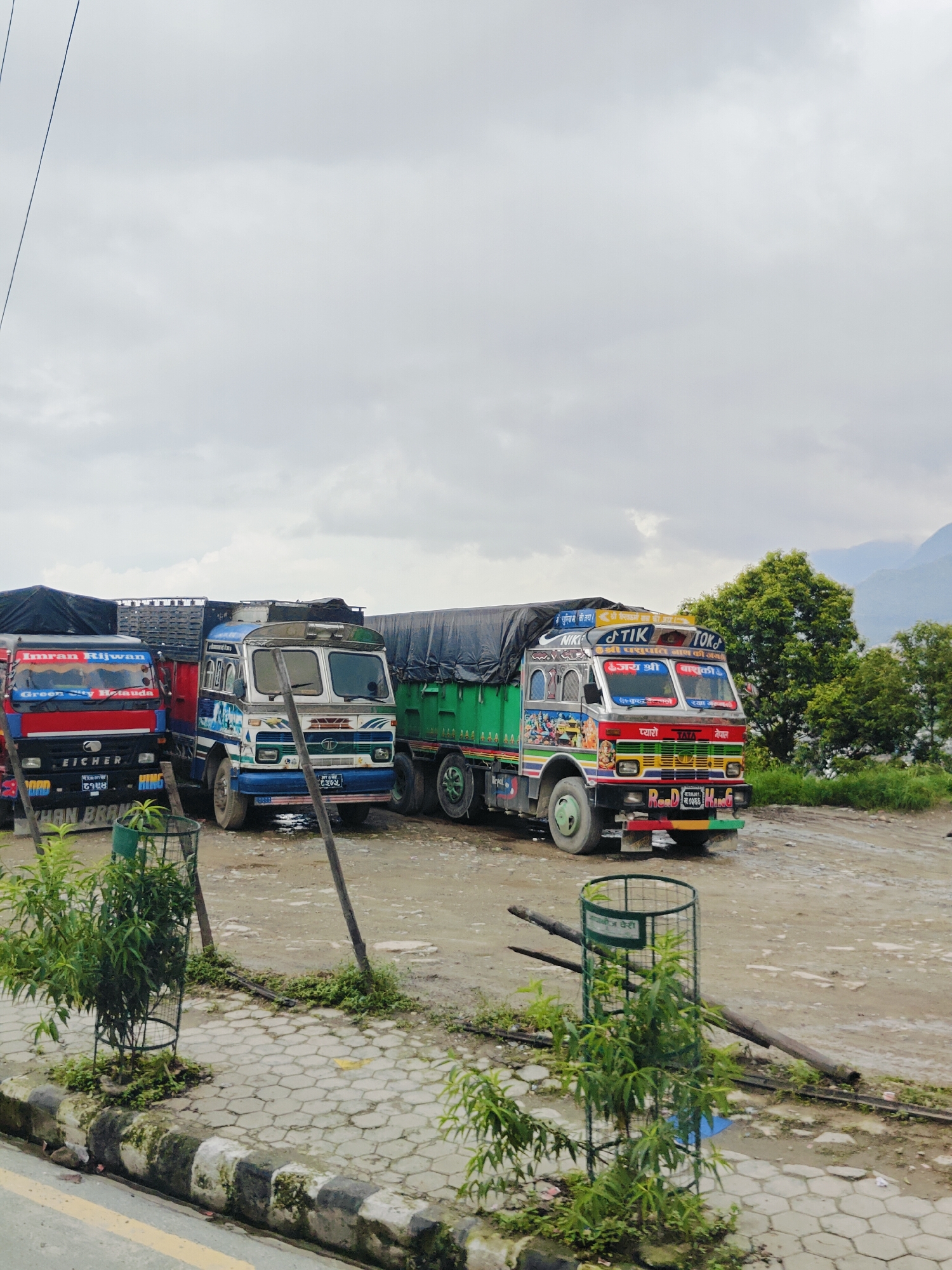 In Chandragiri, Nepal, a line of three colorful, decorated trucks is parked on a dirt and gravel lot. The truck on the far left is red and black, with "Imran Rijwan" and "Green City Hetauda" written on its windshield. The middle truck is white and blue, with "TATA" visible on the front. The truck on the right is predominantly green and adorned with vibrant paintings and lettering in Nepali. Trees and lush greenery are visible in the background, with hazy mountains in the distance under a cloudy sky. A paved walkway with a decorative border runs along the foreground, where several small trees in protective cages are planted. The ground around the trucks appears damp, suggesting recent rain.