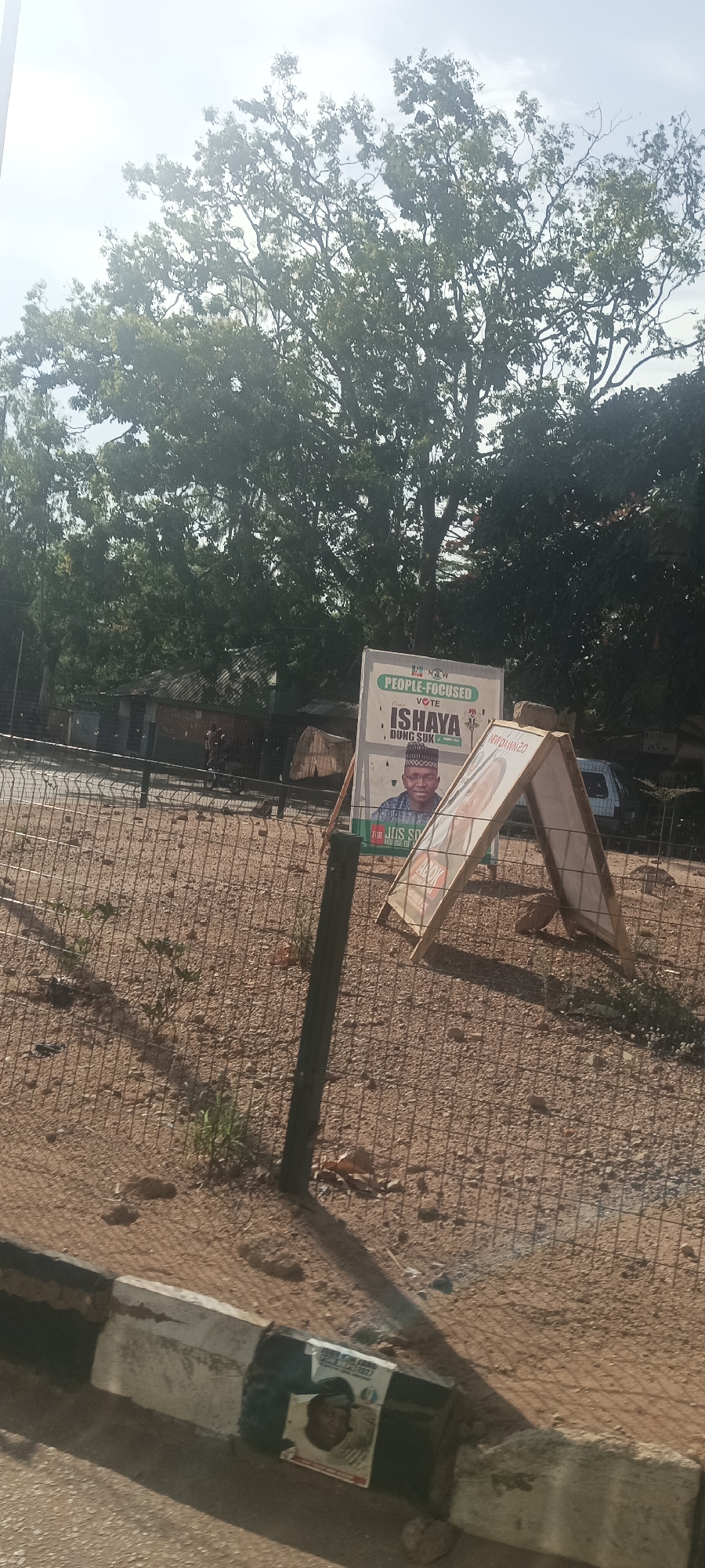 The image captures a street scene in Bukuru, Nigeria. Political campaign posters are prominent. The primary poster features a man named Ishaya Dung Suk, associated with the "New Dawn" movement, identified as "People-Focused." A smaller poster with a portrait of another man is affixed to a roadside concrete barrier. The environment includes a dirt and gravel area enclosed by a fence, with trees visible in the background. A vehicle is also visible in the background. A secondary A-frame poster is next to the "ISHAYA" poster. There are no individuals, animals, or interactions depicted in the scene.