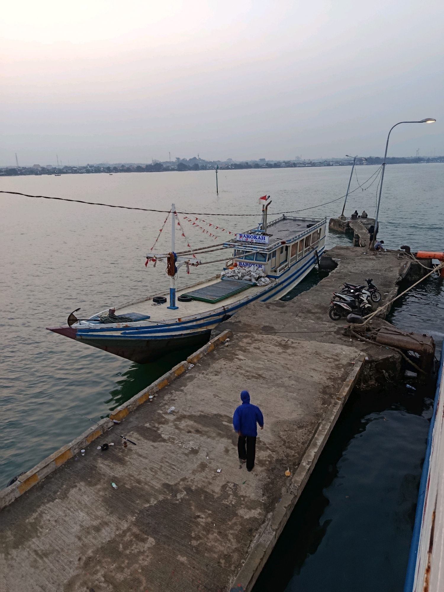 The image depicts a harbor scene in Jepara, Indonesia, likely during the early morning or late afternoon given the soft, diffused light and muted colors. A large, multi-decked passenger ferry, named "BAROKAH," is docked alongside a concrete pier. The ferry is painted white with blue trim and has red and white triangular flags strung along its upper decks. Several people are visible on the pier, including one individual in a blue hooded jacket walking away from the camera on the pier, and a few others gathered near a lamppost at the end of the pier. Two motorcycles are parked on the pier, adjacent to the ferry. The water is calm, with a slight ripple, and the distant shoreline shows a collection of buildings and structures indicative of a coastal town. The pier itself is weathered concrete, with some debris scattered on its surface, including plastic cups and a plastic bottle. Fishing rods are also visible lying on the pier near the water's edge. The overall atmosphere is one of a working harbor, with a hint of the approaching or receding day.