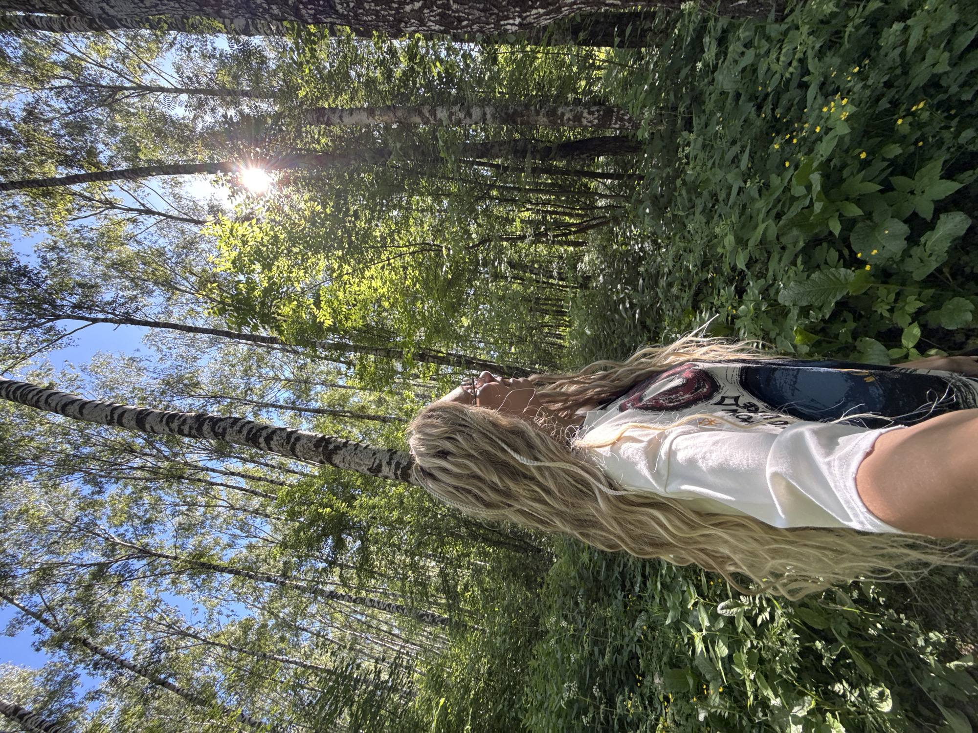 A bright, sunny, eye-level outdoor shot depicts a dense forest scene in Rusanovo, Russia. The primary subject is an individual with long, wavy, light blonde hair, positioned in profile on the left side of the frame, facing right. The individual wears a light-colored long-sleeved shirt featuring a graphic design with dark blue, red, and white elements. Eyeglasses with light frames are visible on the person’s profile. The right arm extends into the lower right foreground, suggesting the person is holding a camera or smartphone to take the photograph.
The surrounding environment is a deciduous forest, characterized by numerous tall, slender trees with light-colored bark and dark horizontal stripes, consistent with birch trees. The leaves on the trees are predominantly light green, indicating spring or early summer foliage. Patches of clear blue sky are visible through the tree canopy. Intense sunlight emanates from the upper right quadrant of the frame, creating a distinct sunburst effect through the leaves. The forest floor is thickly covered with lush green undergrowth, including various herbaceous plants and scattered small yellow wildflowers, particularly in the lower left and right areas. The scene conveys a sense of depth into the woodland.