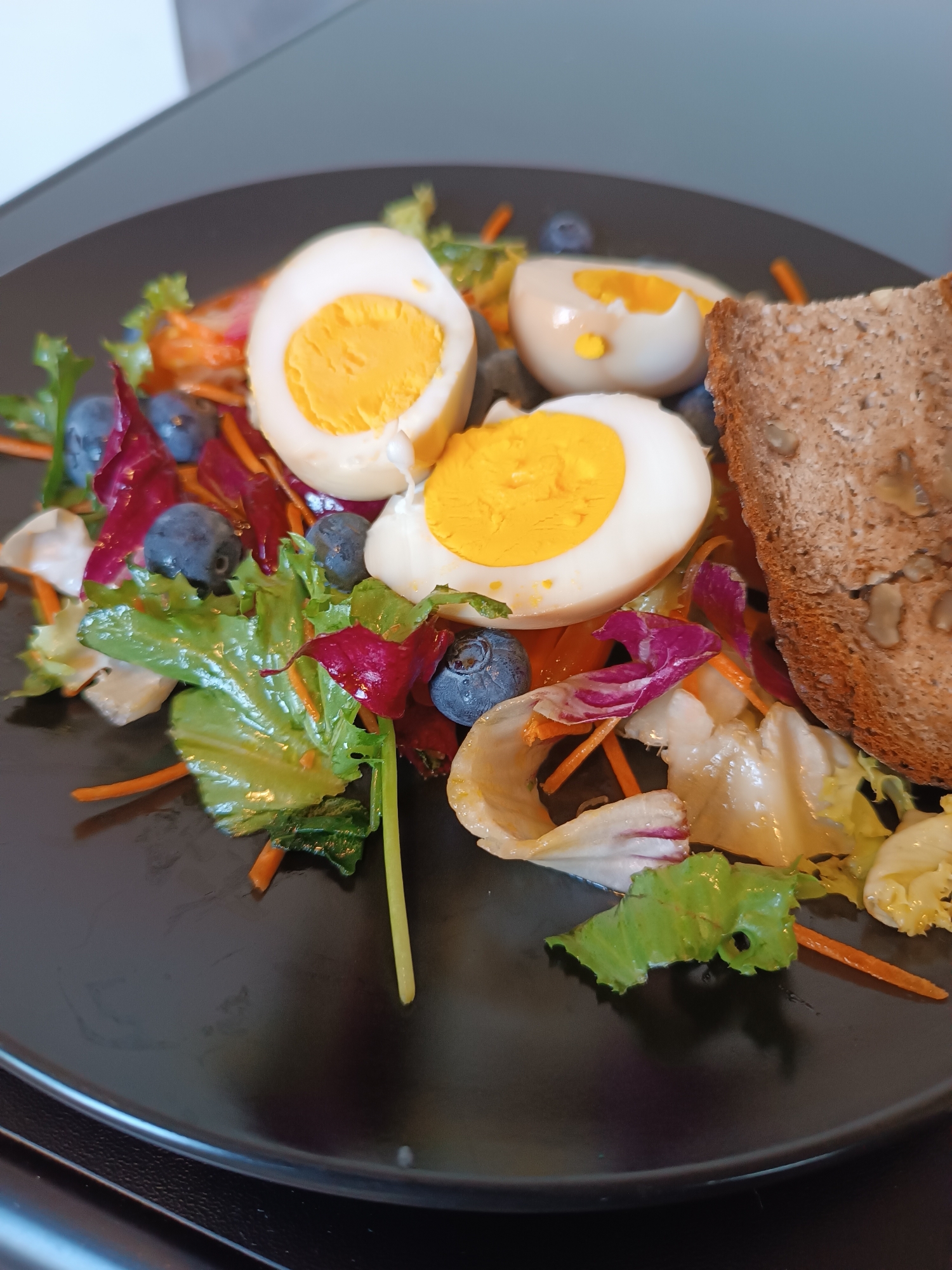 A close-up, eye-level shot shows a black plate with a salad and a slice of bread. The salad consists of mixed greens, shredded carrots, blueberries, and sliced hard-boiled eggs. A single slice of dark, seedy bread is positioned on the right side of the plate. The background is out of focus, showing a gray surface and a white object. The lighting is natural and even. There are no people visible in the image. This scene depicts a meal, likely breakfast or lunch, served on a plate. It is impossible to determine the location within the city of Viadana, Italy, from the provided image.