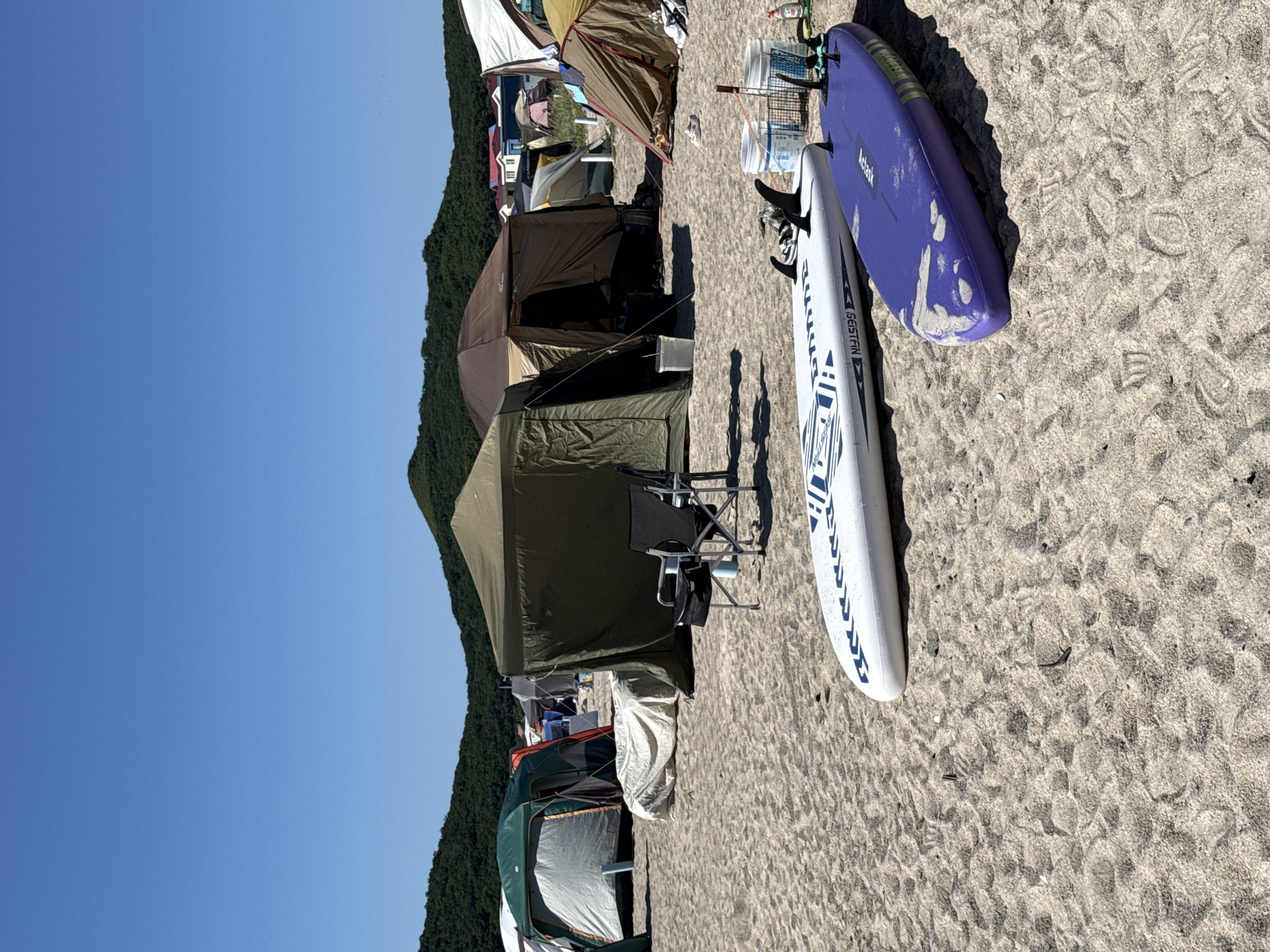 A sunlit, sandy beach campsite in Nakhodka, Russia, is depicted under a clear blue sky. The scene features multiple tents of various sizes and colors, suggesting a group camping expedition. In the foreground, two paddleboards are laid out on the sand; one is white with blue lettering and geometric patterns, and the other is purple with some visible scuff marks. Behind the paddleboards, several white buckets and a metal basket are present, possibly used for storing or transporting items. A black camping chair is positioned near a large, olive-green tent. The backdrop includes rolling, tree-covered hills and a glimpse of houses with red roofs in the distance. The sand is textured with footprints and ripples.