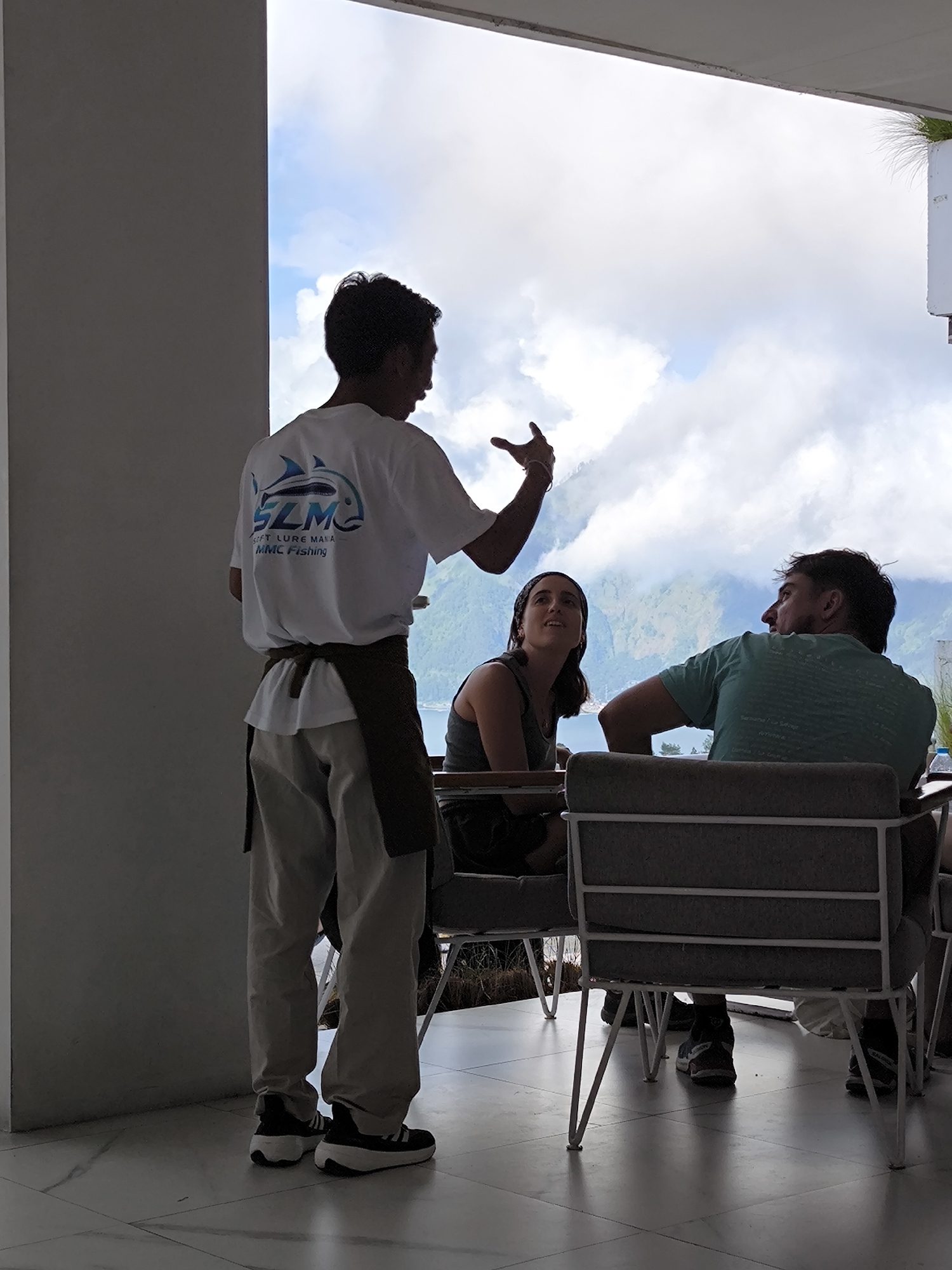The image captures a scene at a restaurant in Kintamani, Indonesia, overlooking a lake and mountain vista under a cloudy sky. A waiter stands, facing the couple sitting at a table. He is wearing a white t-shirt with a logo for "SLM MMC Fishing" on the back, light-colored pants, and black-and-white sneakers, with a brown apron tied around his waist. He is gesturing with his right hand while addressing the couple.
The couple is seated at a table with grey chairs. The woman is wearing a dark-colored top and looking up at the waiter. The man is wearing a green t-shirt and turning his head towards the waiter. The table and chairs are situated near a large opening offering an expansive view of the lake and mountains beyond. The interior of the restaurant features white marble flooring and a large white pillar on the left side.