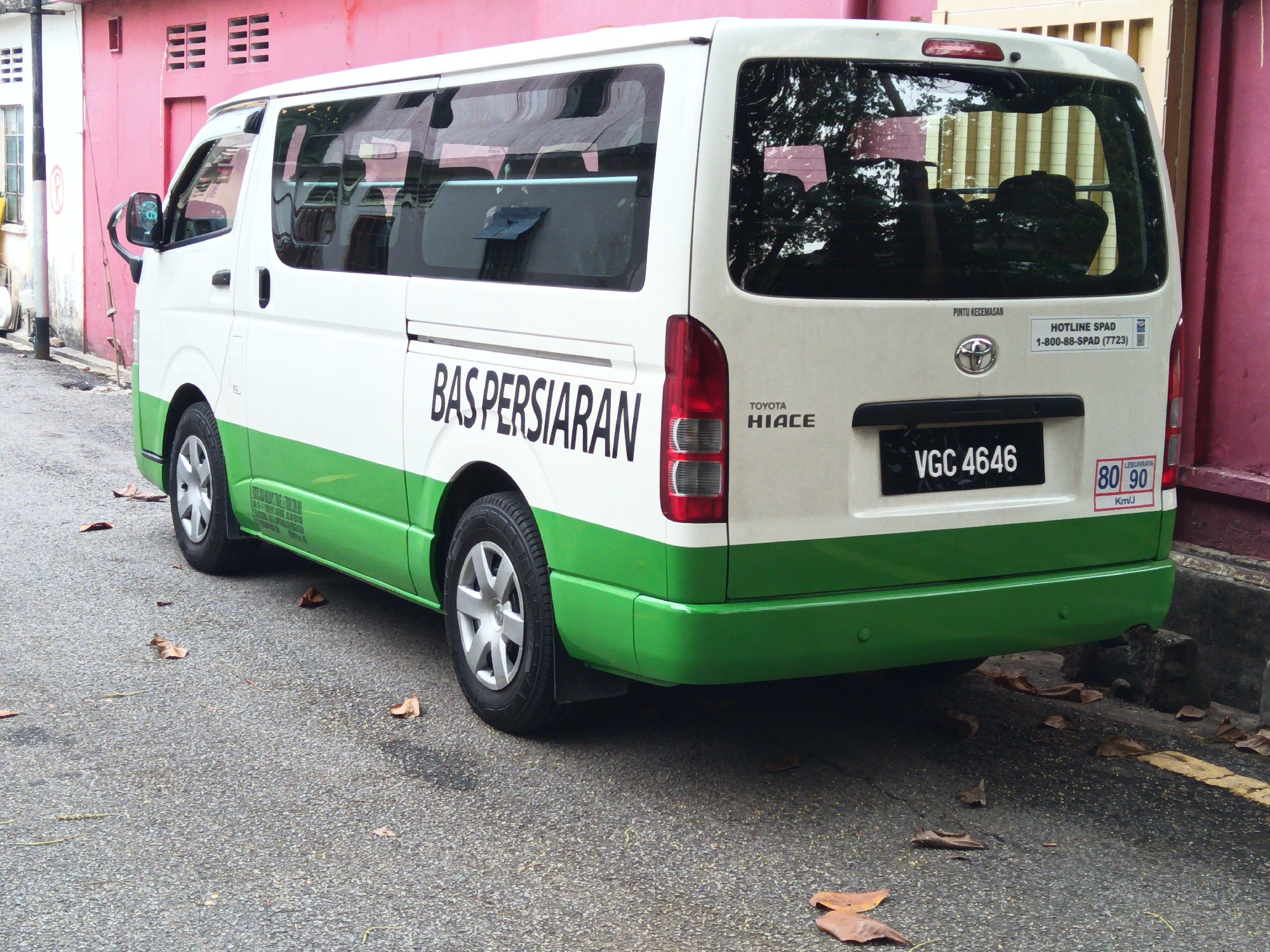 A white and green Toyota Hiace van is parked on a paved road in Kuala Lumpur, Malaysia. The van is white on top with a green lower body. On the side of the van, the words "BAS PERSIARAN" are written in large black letters. The license plate of the van reads "VGC 4646". There are various stickers on the rear of the van, including a "HOTLINE SPAD" sticker and two speed limit stickers, "80" and "90". The van is parked next to a pink building with horizontal window slits. The ground is covered with scattered dry leaves. The scene appears to be a residential or commercial street.