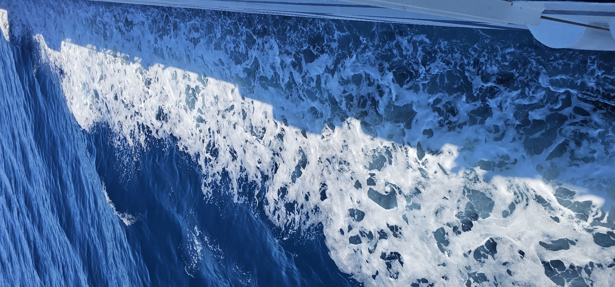 This is a high-angle, vertical shot of a boat's wake on the sea, seen from the perspective of someone on board. The water is a deep blue and frothy white, with the white foam churned up by the boat's passage creating a distinct line separating the calmer water from the turbulent wake. A white railing or side of the boat is visible on the right edge of the frame, casting a long shadow across the water. The scene appears to be set on the sea near the city of Supetar, Croatia.