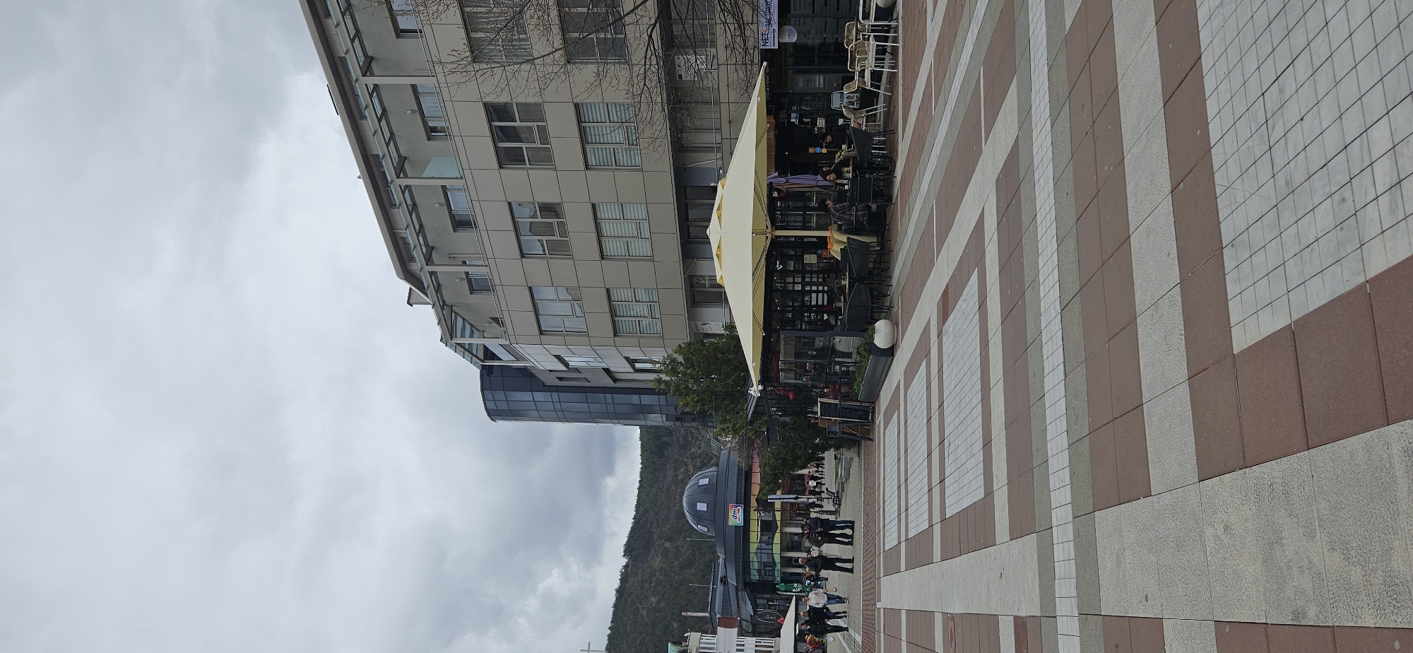 The image shows a street scene in Blagoevgrad, Bulgaria. The perspective is from a low angle, looking towards a row of buildings and a paved square.
**Location:** The scene appears to be in the city center of Blagoevgrad, Bulgaria. A distinctive building with a dome (likely a commercial or municipal building) is visible along with other modern architecture.
**Environment:**
* Overcast sky with grey clouds.
* A paved square with a red and white tiled pattern occupies a large portion of the foreground.
**Subjects/Objects:**
* A multi-story building with a facade of grey panels and windows lines the left side of the image.
* A semi-circular modern building with a dark glass facade is partially visible next to the multi-story building.
* A cafe or restaurant with a yellow canopy is positioned on the square. Tables and chairs are visible underneath the canopy.
* A line of people walking along the edge of the square.
**People:**
* Several people are walking along the street near the buildings.
**Actions/Interactions:**
* People walking on the sidewalk or the square.
* Patrons sitting at the cafe.