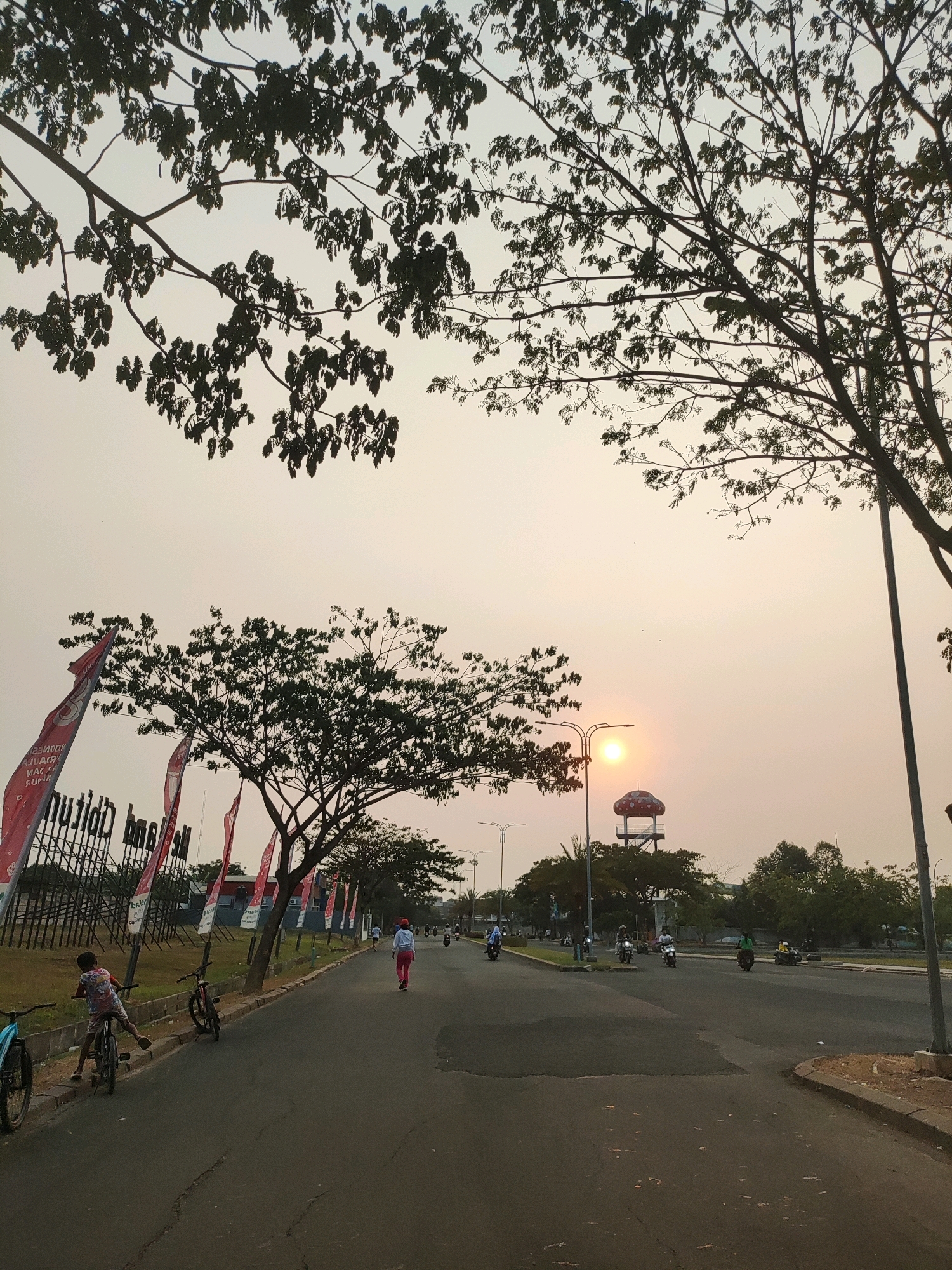 In Telagamurni, Indonesia, a street scene unfolds under a hazy sky at sunset. The sun, a soft orange orb, is low on the horizon, casting a warm glow. Trees with silhouetted branches frame the upper portion of the image. A road curves through the scene, with several people and vehicles present. On the left side of the road, a young child on a bicycle is partially visible, with another bicycle next to them. Further down the road, a person in a red hat and pink pants is jogging. Several motorcycles are also on the road, moving in the same direction as the jogger. In the background, a water tower with a red, mushroom-shaped cap stands out. Red flags with white text are planted along the side of the road, and a fence encloses an area behind them. The environment appears to be an urban or suburban area, possibly a park or a residential development, given the presence of recreational activities and the organized landscaping.