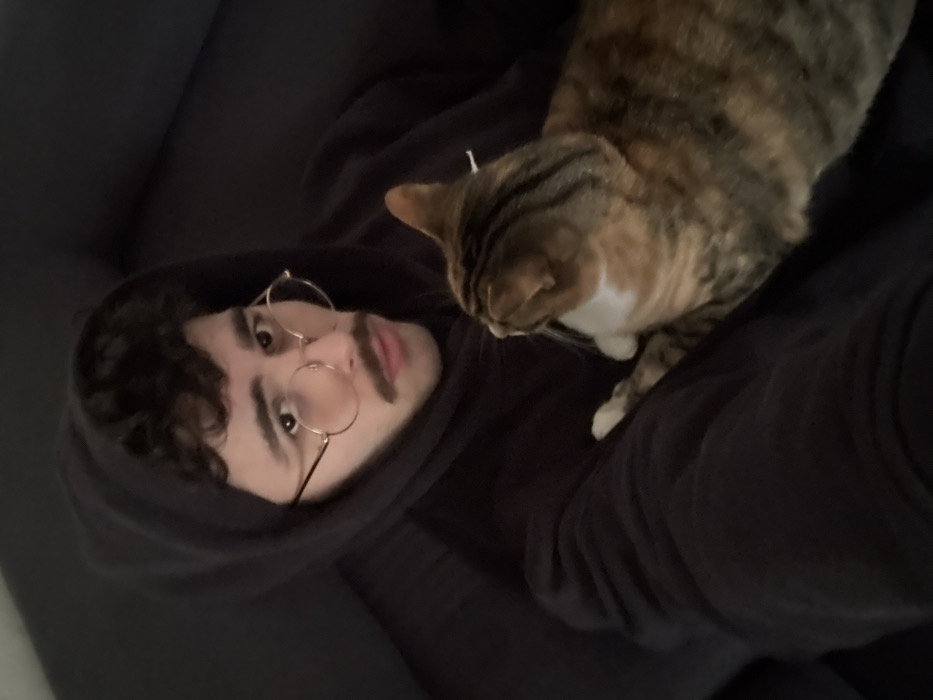 An indoor scene depicts a young adult male with dark, curly hair, a mustache, and round, gold-rimmed glasses. He is wearing a dark hooded sweatshirt with the hood up, and is seated against a dark upholstered surface, possibly a couch or armchair. He looks directly into the camera with a neutral expression. A domestic short-haired cat with a calico or tortoiseshell coat (brown, orange, and white patches) is resting on his lap or chest, facing away from the camera and towards the viewer's right. The cat's white chest and front paws are visible. The specific location in Rennes, France, cannot be determined from the visual information provided in the image.