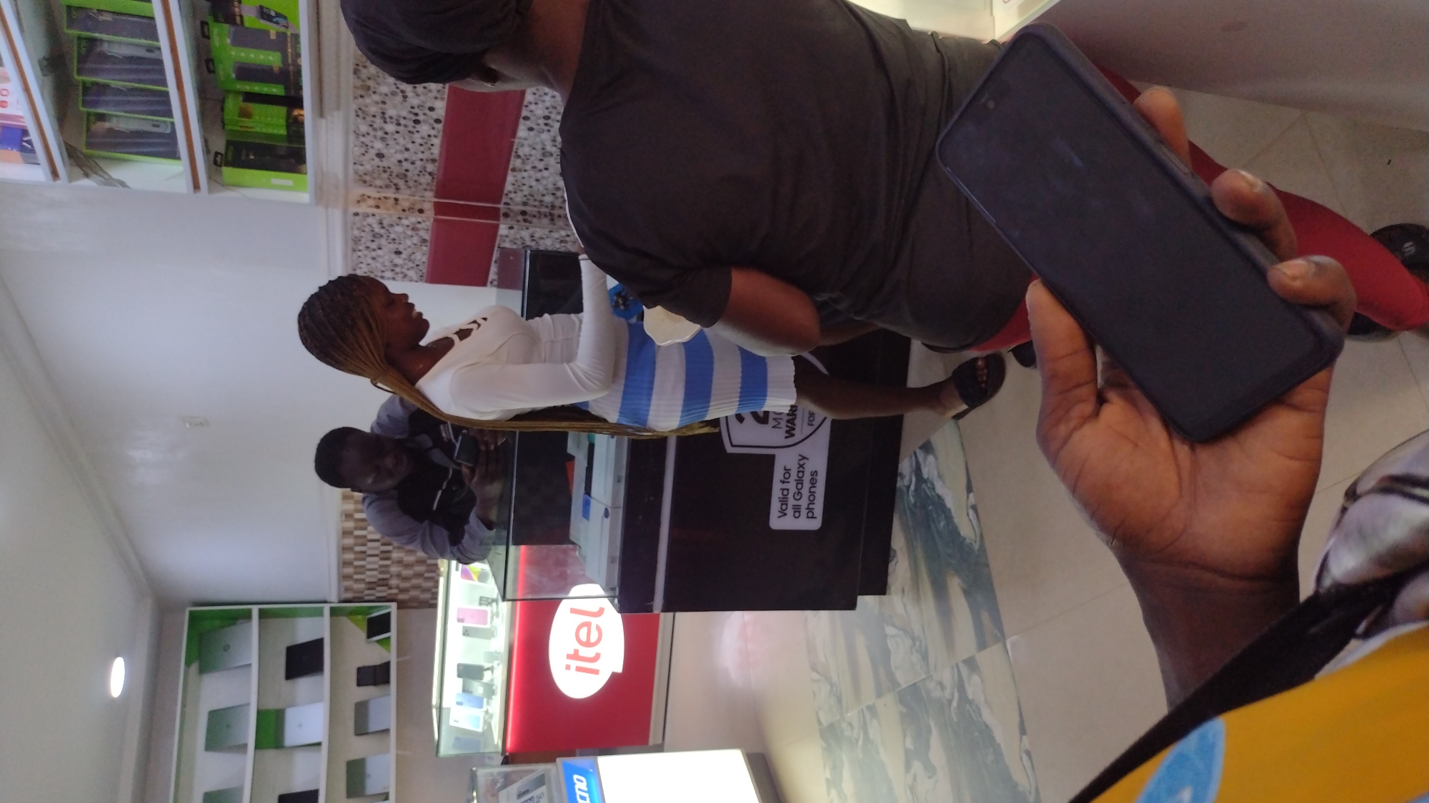 The image captures a scene within a retail setting, likely a mobile phone store in Jos, Nigeria.
Several individuals are present: a woman with blonde braids wearing a white long-sleeved shirt and a blue and white striped skirt stands near a glass display case. Another man is seen in front of the glass display case and is holding a phone. Partially visible is a person wearing a gray shirt. In the foreground, a hand holds a black smartphone; this is presumably the point of view of the image taker.
The environment includes a display case with "itel" branding. The sign on the display case reads "Valid for all Galaxy phones WAR." Shelves filled with mobile phones are visible in the background. The floor is tiled.