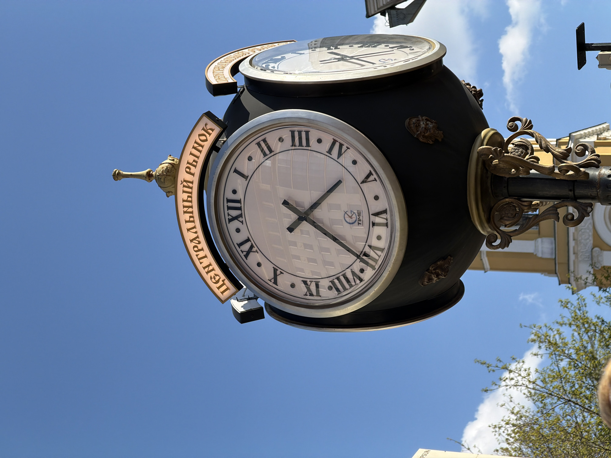A low-angle, outdoor shot depicts a decorative street clock against a bright blue sky with scattered clouds. The clock, likely situated in Moscow, Russia (based on the Cyrillic inscription), features two faces encased in a dark, possibly black, spherical housing. Both clock faces are white with black Roman numerals and hands, although they may display slightly different times. The bottom clock face is partially obscured, but the hands appear to indicate roughly 5:45. The clock is mounted on an ornate, gold-colored post, adorned with decorative scrollwork and potentially stylized faces or carvings. A sign, inscribed in Cyrillic, curves around the left side of the clock; the words "ЦЕНТРАЛЬНЫЙ РЫНОК" (Central Market) are visible. In the background, a building with yellow walls and white trim is partially visible, and the branches of a tree extend into the frame on the lower right side. The sky occupies most of the upper portion of the image, highlighting the clock as the central subject.