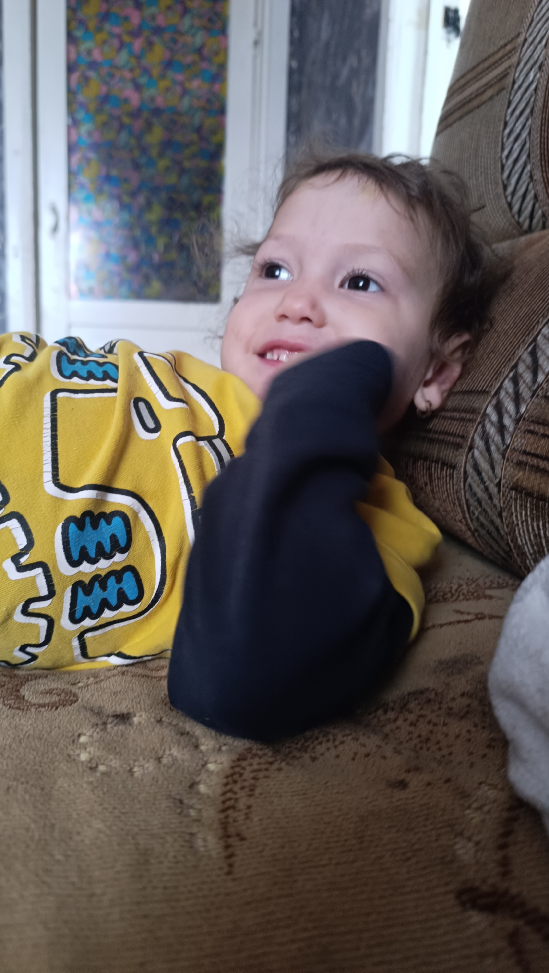 A close-up, waist-up shot shows a young child lying on their back on a brown, textured couch. The child, who appears to be a toddler, is wearing a bright yellow t-shirt with a black and blue graphic design on the front. Their head is turned to the left, and their eyes are looking upwards. The child's right hand, covered in a black sock, is held up near their face. The background is blurry, but a window with colorful stained glass is visible to the left, and a portion of a brown upholstered chair is seen to the right. The lighting suggests an indoor environment, possibly a living room. The image does not contain any information about the city of Karsha, Russia.