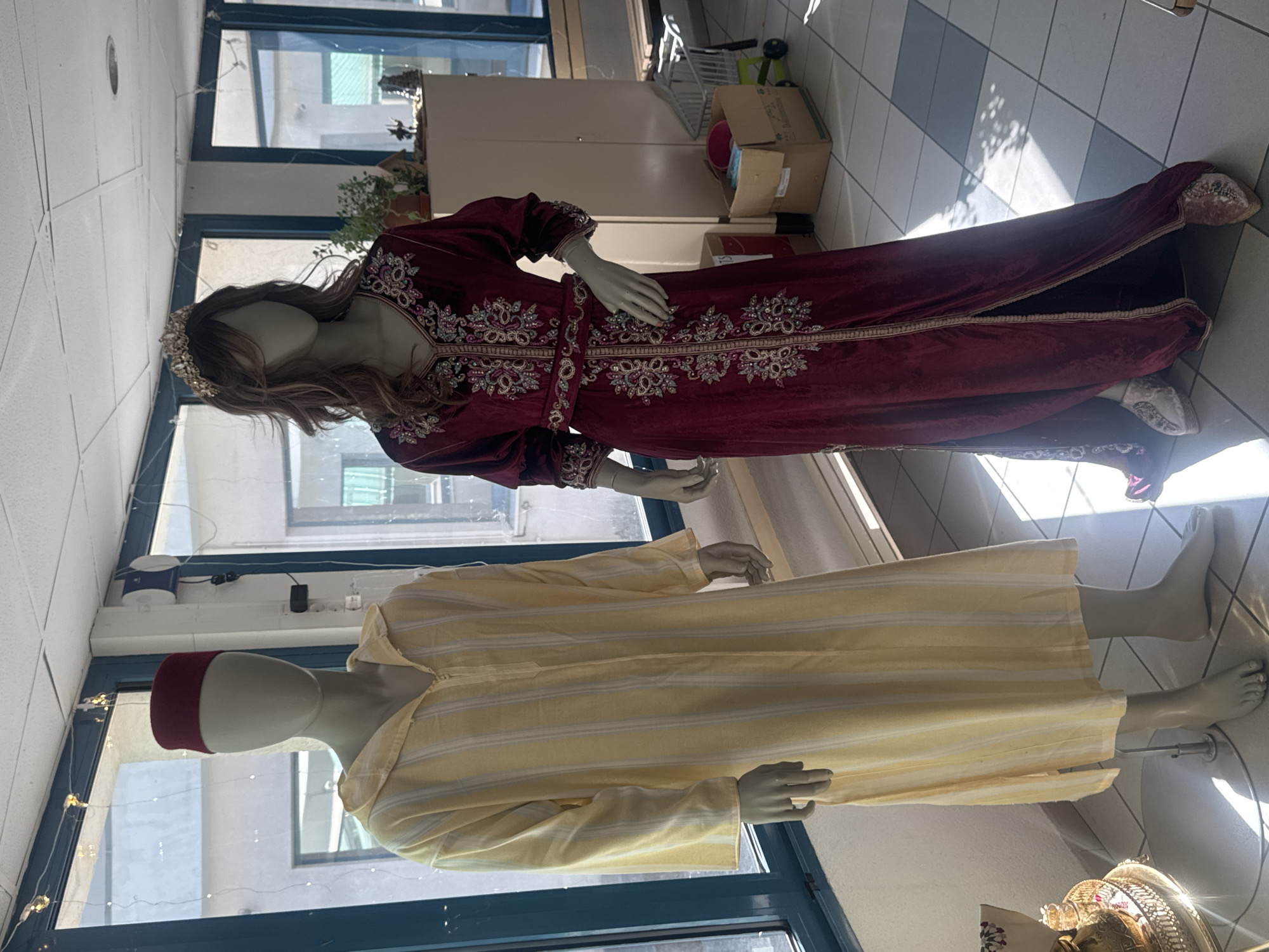 The image shows two mannequins in what appears to be a retail space, possibly a boutique or shop, located in Saint-Claude, France.
The mannequin on the left is wearing a light yellow, striped djellaba, a traditional long robe, and a dark red fez hat. The djellaba has a zipper closure down the front and a hood.
The mannequin on the right is dressed in a deep red velvet caftan adorned with intricate silver and purple embroidery. It features a V-neckline and long sleeves, and is cinched at the waist with a matching embroidered belt. This mannequin also wears a decorative crown and what appear to be embellished shoes. Its hair is a medium brown with wavy texture.
The background suggests an indoor setting with large windows that let in natural light, creating distinct shadows on the tiled floor. Behind the mannequins, there are shelves with various items, including plants and decorative objects. Further in the background, a white cabinet and a shopping cart are visible. The overall lighting is bright, with sunlight streaming through the windows. The floor is tiled in a checkerboard pattern of light and dark grey squares.
