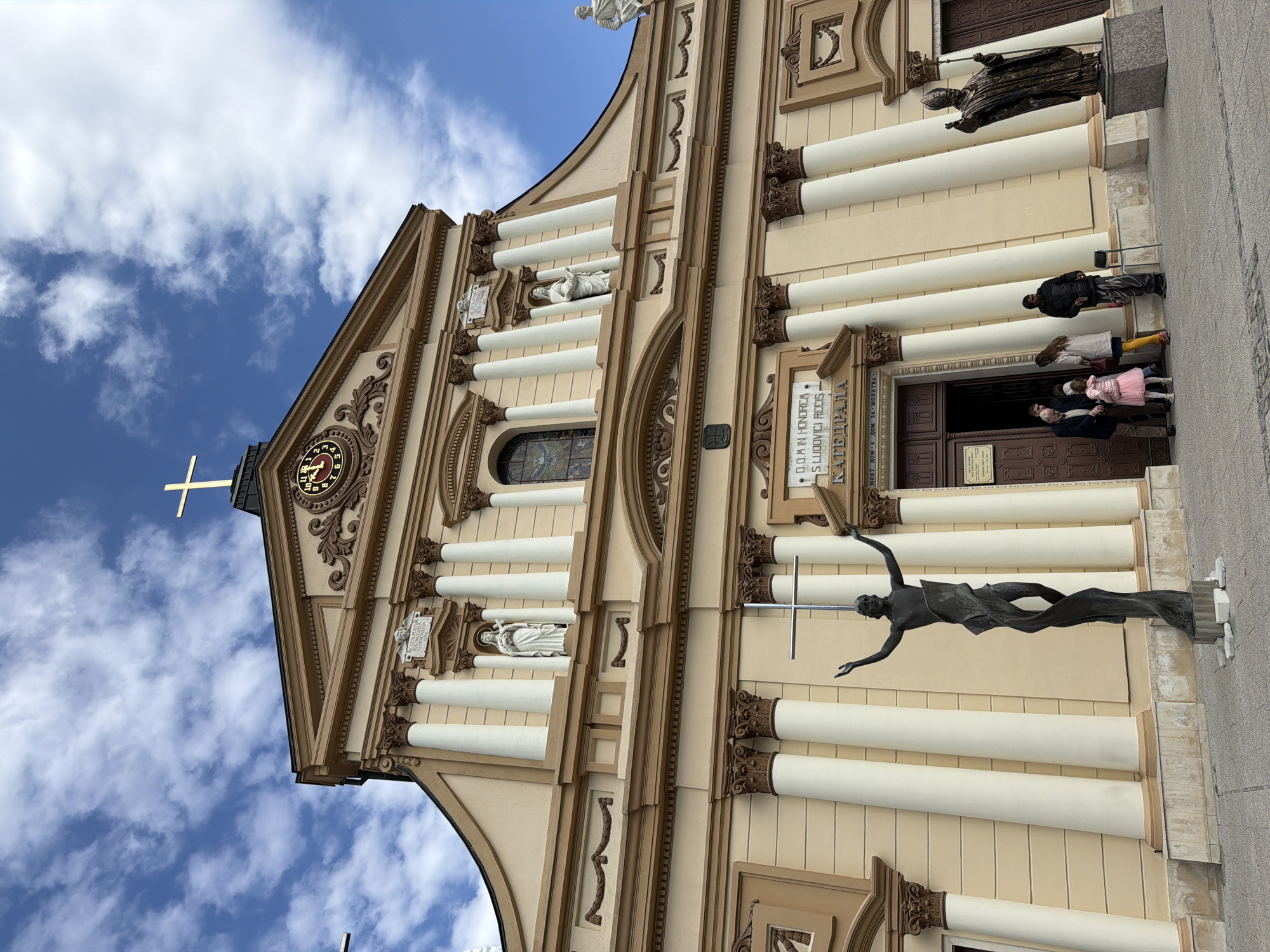 The image depicts the exterior facade of the Cathedral of St. Louis in Plovdiv, Bulgaria. The cathedral features a classical architectural style with columns, statues, and decorative elements. A prominent sculpture of a figure holding a cross is in the foreground. Several people are standing near the entrance of the building. The sky is visible in the background with blue color and white clouds.