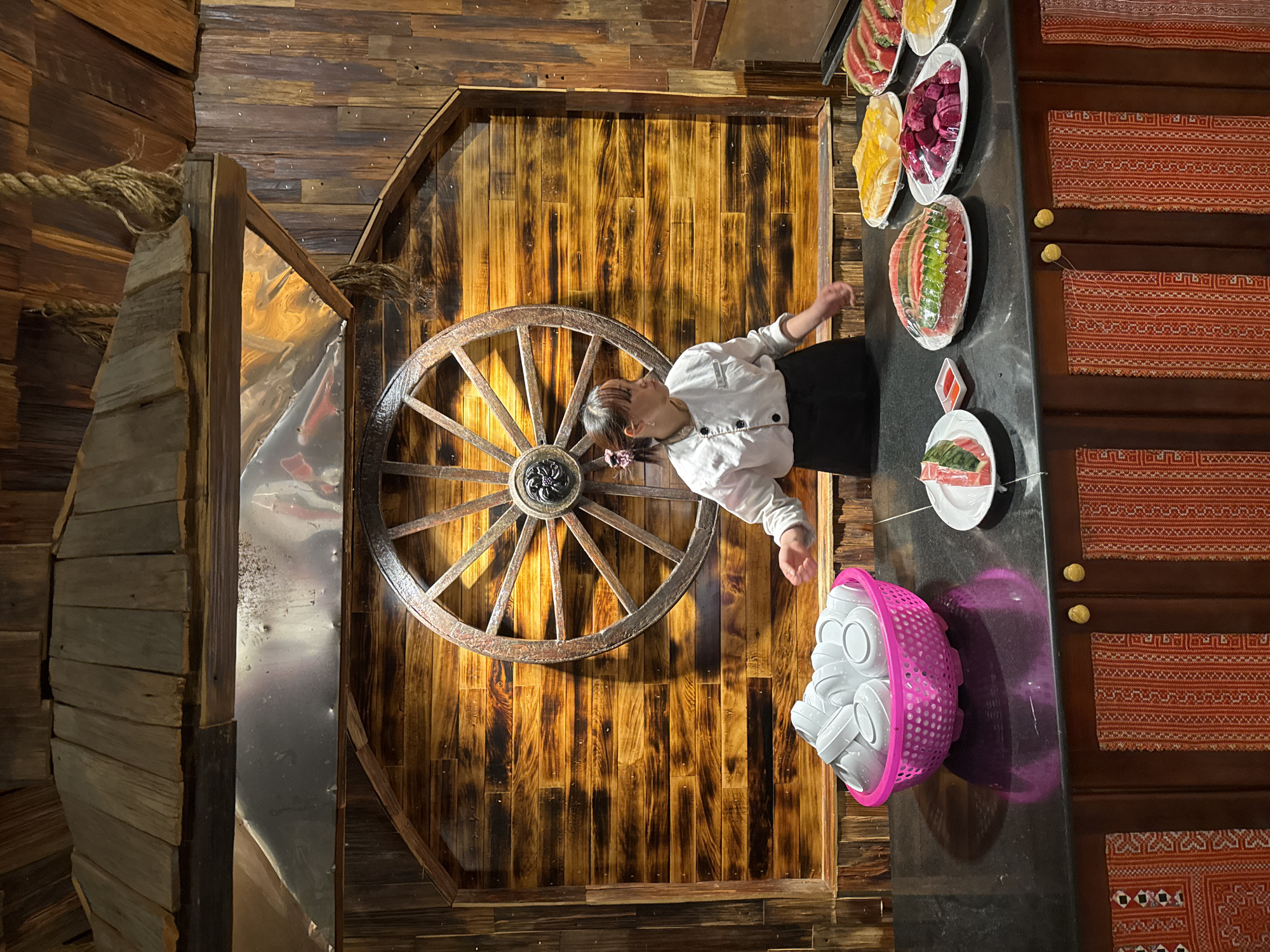 A horizontally-oriented photograph depicts the interior of a venue in Sa Pa Ward, Vietnam, featuring a young person in a chef's coat standing next to a table laden with food and cups, set against a backdrop of rustic wooden walls and decorative elements. The venue is identifiable by its unique design features.
The person is dressed in a white chef's coat and dark pants, positioned near a black counter top. A variety of dishes, likely part of a buffet, are arranged along the counter, containing sliced fruits and vegetables, and what appears to be prepared meat dishes. A pink plastic basket filled with numerous white cups sits nearby.
The walls are composed of textured, dark-stained wood, suggesting a rustic or country-themed establishment. The room's wall is decorated with a large wooden wagon wheel adding to the rural aesthetic. The setting includes a section of a cabin-like or rustic structure to the left, made of roughly-hewn planks and held together with rope. To the right of the counter is a wooden cabinet with decorative, patterned textiles covering its front.
