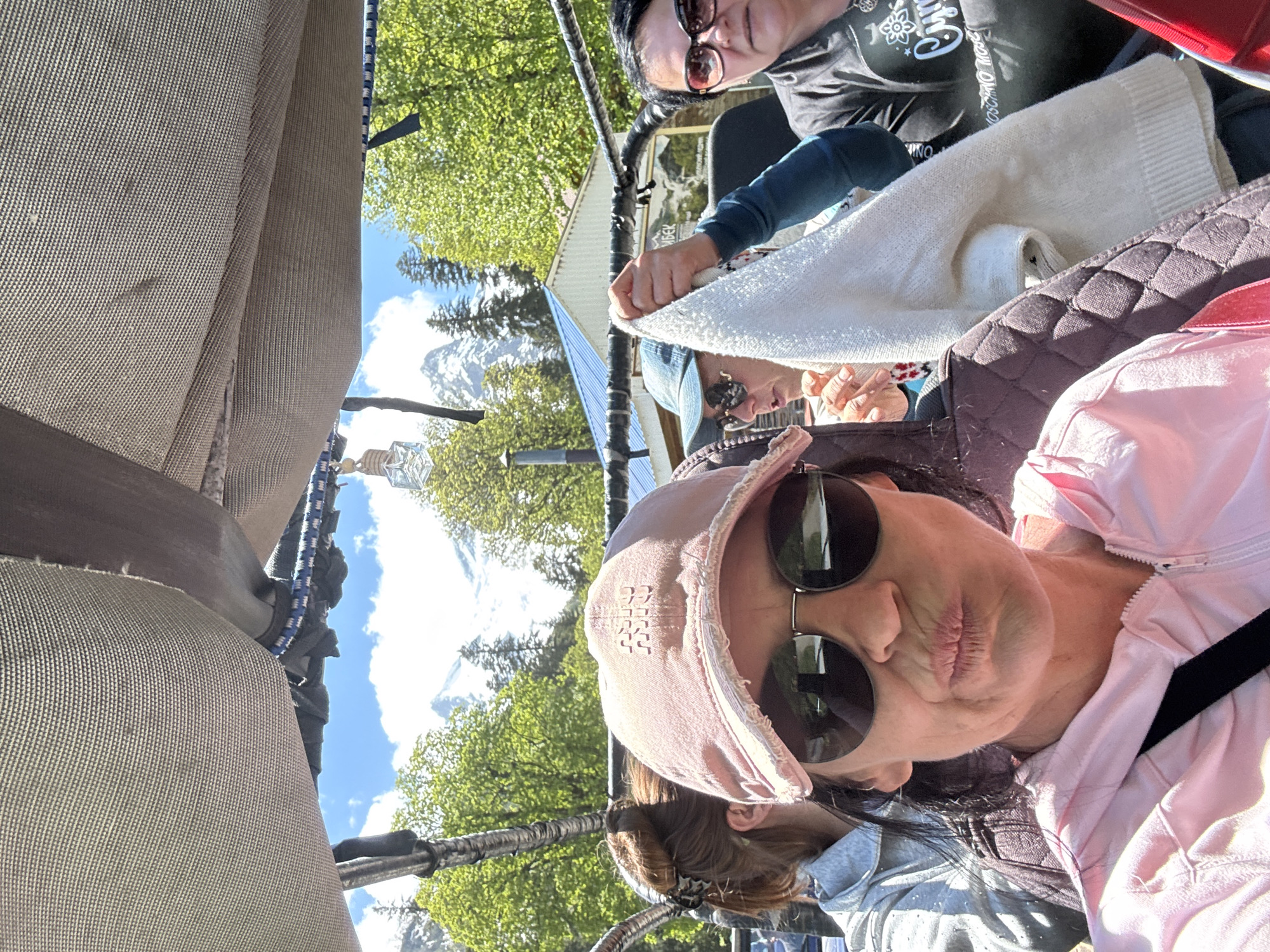 The image captures a selfie from a partial interior perspective, likely within an open-air vehicle or cabin, on a bright, sunny day in the mountainous region of Верхне-Тебердинское сельское поселение, Russia.
In the foreground, a woman with dark hair and a pink, distressed baseball cap bearing the Cyrillic text "МИШУ" (MISHU) on the front is taking the photo. She is wearing dark aviator-style sunglasses and a pink hooded jacket, making a puckered lip expression. A black strap is visible across her chest.
Behind her on the left, partially visible, is another woman with light brown hair tied in a bun with a dark claw clip, wearing a light-colored jacket. To the right, a third woman wearing a blue baseball cap and sunglasses holds a folded white knitted sweater or blanket with both hands. Further to the right, a fourth woman, partially visible, wears dark sunglasses and a dark t-shirt with white Cyrillic lettering, including "МОС" (MOS).
The immediate overhead structure is covered with a beige, woven fabric canopy, from which a blue-and-white striped bungee cord and a small, star-shaped glass bottle hang. Metallic, ribbed tubing forms part of the structure around the occupants.
The background reveals a vibrant natural landscape under a clear blue sky with scattered white clouds. Lush green deciduous trees and some evergreen trees are prominent. In the distance, snow-capped mountains rise majestically. A building with a blue corrugated metal roof and a chimney is visible in the mid-ground. Adjacent to the building, a sign with Cyrillic text, including "ТЕБЕРДИНО-КУР..." (likely Teberdino-Kurort or similar, indicating a resort area), is partially visible, confirming the location within the Teberda region of Russia.
