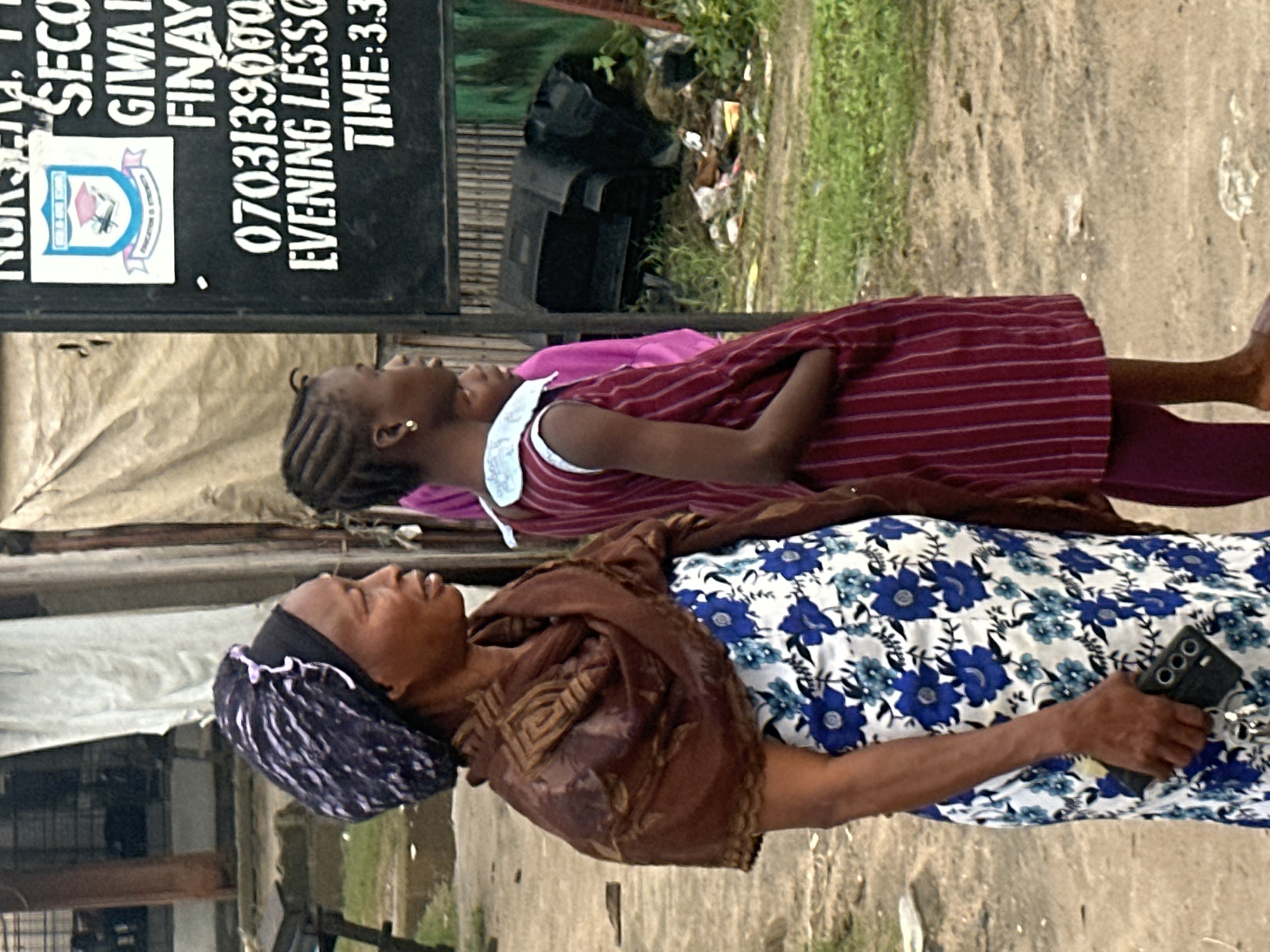 The image depicts a street scene in Badagry, Nigeria, focusing on two individuals. In the foreground, an older woman wears a dark blue headscarf with white streaks and a brown patterned shawl draped over a white dress with blue floral motifs. She is holding a black mobile phone in her right hand. To her right stands a younger girl in a maroon and white striped dress, her hair in braids, and wearing a gold earring. She has her right hand in her pocket. Behind them, a portion of a black chalkboard sign is visible, displaying text including a phone number "0703139000", "EVENING LESSONS", and "TIME: 3.3". The environment appears to be a street with dirt ground and some vegetation. In the background, structures made of wood and possibly corrugated metal are present, along with discarded items and debris, suggesting an urban setting that may be undergoing development or experiencing challenges with waste management.