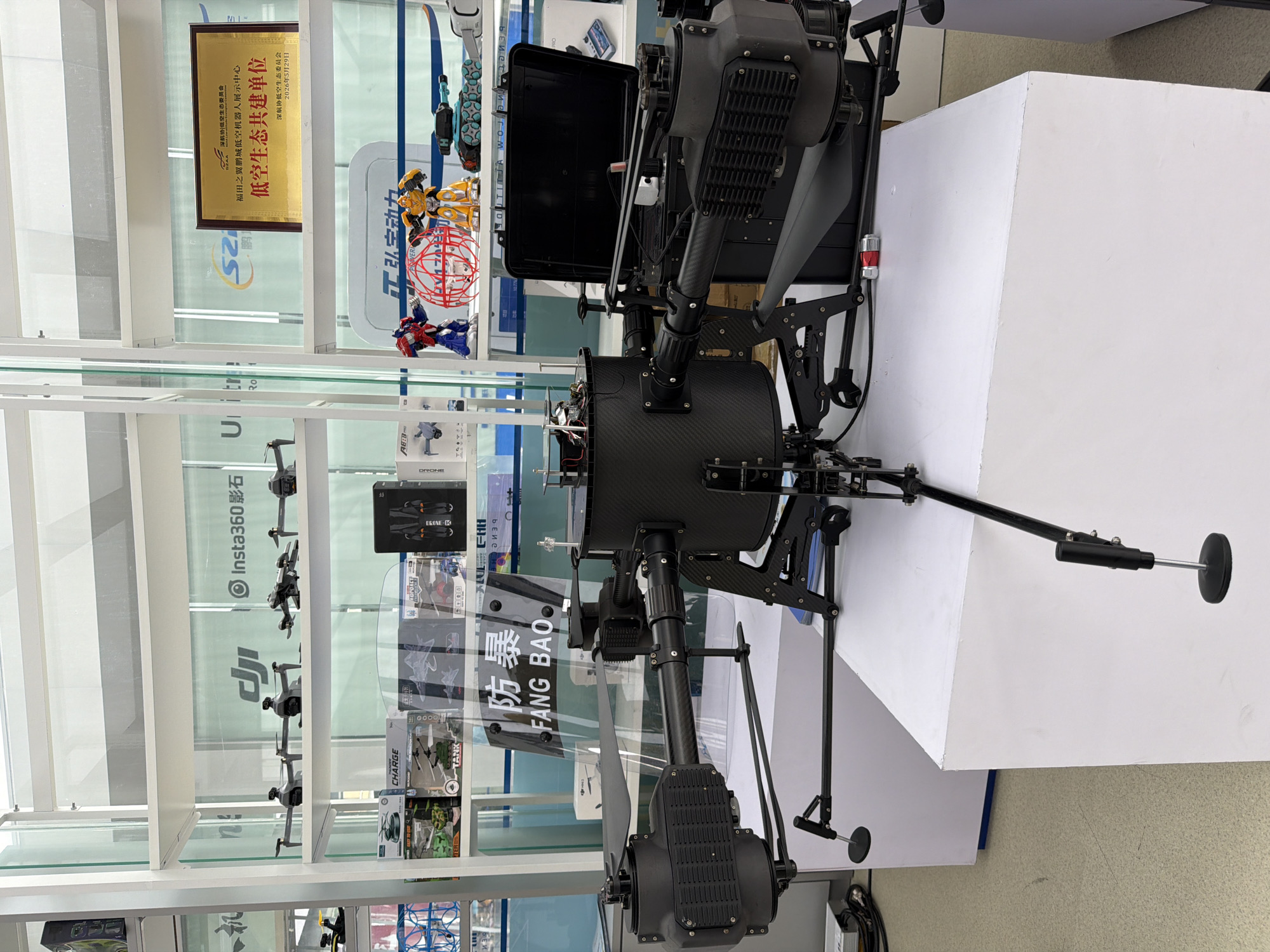 In the Futian District of China, a large, black drone with multiple rotors is displayed prominently on a white pedestal. The drone features a central cylindrical body and four extended arms, each equipped with a rotor assembly. A clear shield with the Chinese characters "防暴 FANG BAO" is positioned in front of the drone. Behind the drone, a black case is visible. The drone is housed in a brightly lit exhibition space with white shelving units. These shelves display various smaller drones, toy robots resembling Optimus Prime and Bumblebee, and other electronic devices. A gold plaque is mounted on the wall behind the shelves, indicating an award or recognition for "低空生态共建单位" (Low Altitude Ecological Co-construction Unit). The environment appears to be a commercial or exhibition setting, possibly a drone or technology store, given the variety of products on display.