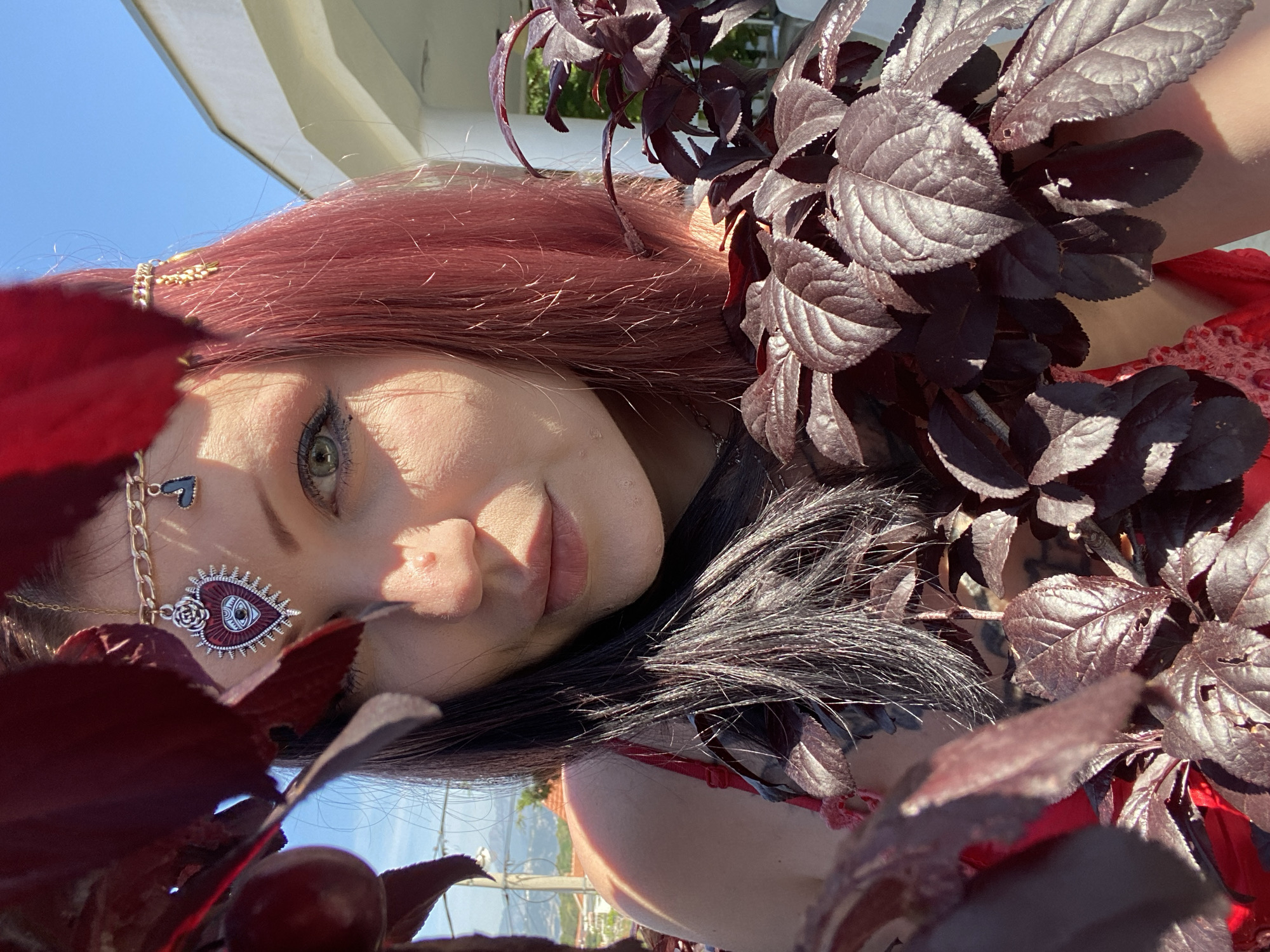 A portrait-style shot captures a young woman with reddish-brown hair and fair skin, positioned outdoors in Kunje, Montenegro. She is looking directly at the camera with a calm, neutral expression, a slight smile playing on her lips, and her green eyes are prominent.
The woman's head is tilted, and her face is partly framed and partially obscured by numerous dark, deep red-purple leaves belonging to a nearby plant or branch. Small, dark red berries are also visible among these leaves in the foreground. Sunlight highlights the textures of the leaves and parts of her hair.
She is adorned with intricate accessories. A delicate gold-colored chain with a small heart charm is visible near her temple. Below her eye, a larger, ornate pendant hangs, featuring an 'evil eye' motif within a red and silver heart design, embellished with rose-like details. Her makeup includes dark eyeliner and mascara, emphasizing her eyes.
A red garment, possibly a top or dress with a subtle lace pattern, is visible on her shoulder and chest. The background features a clear blue sky and a white architectural structure, suggesting a bright, sunny daytime setting. The overall composition creates an ethereal and somewhat mysterious mood, with the dark leaves contrasting with her skin tone and the bright sky. No visible text is present in the image.