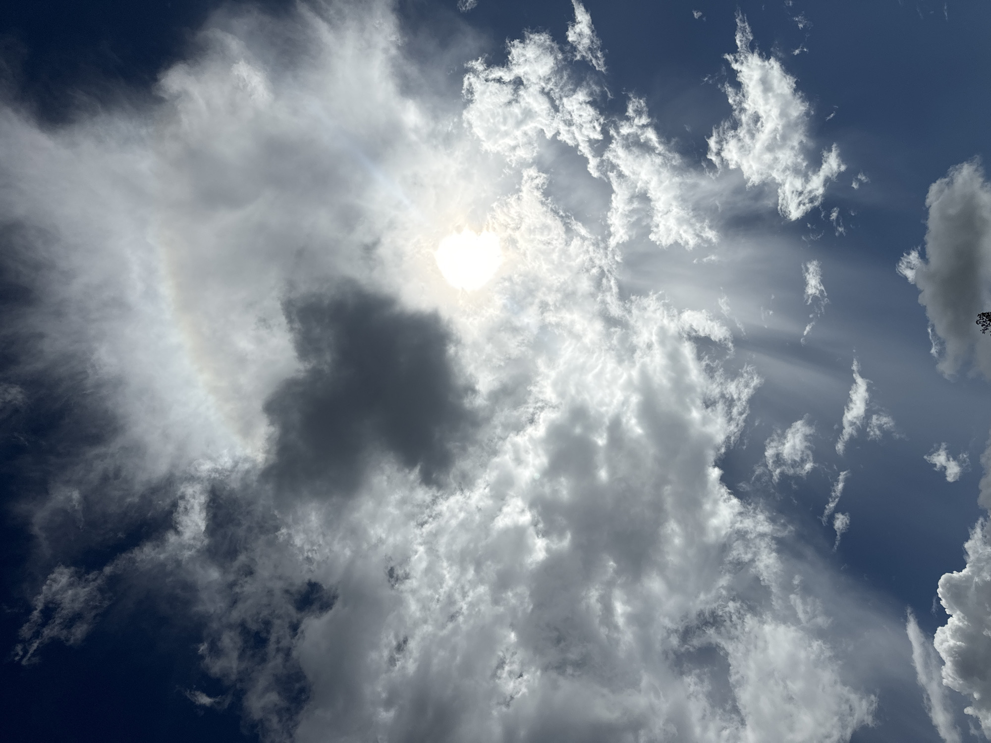 The image is a low-angle shot of the sky in Mũi Né, Vietnam. The sky is mostly blue, with a few scattered clouds of varying sizes and shapes. The sun is visible in the center of the image, partially obscured by a large cloud. A full halo around the sun can be seen. The image does not contain any people, actions, or interactions.
