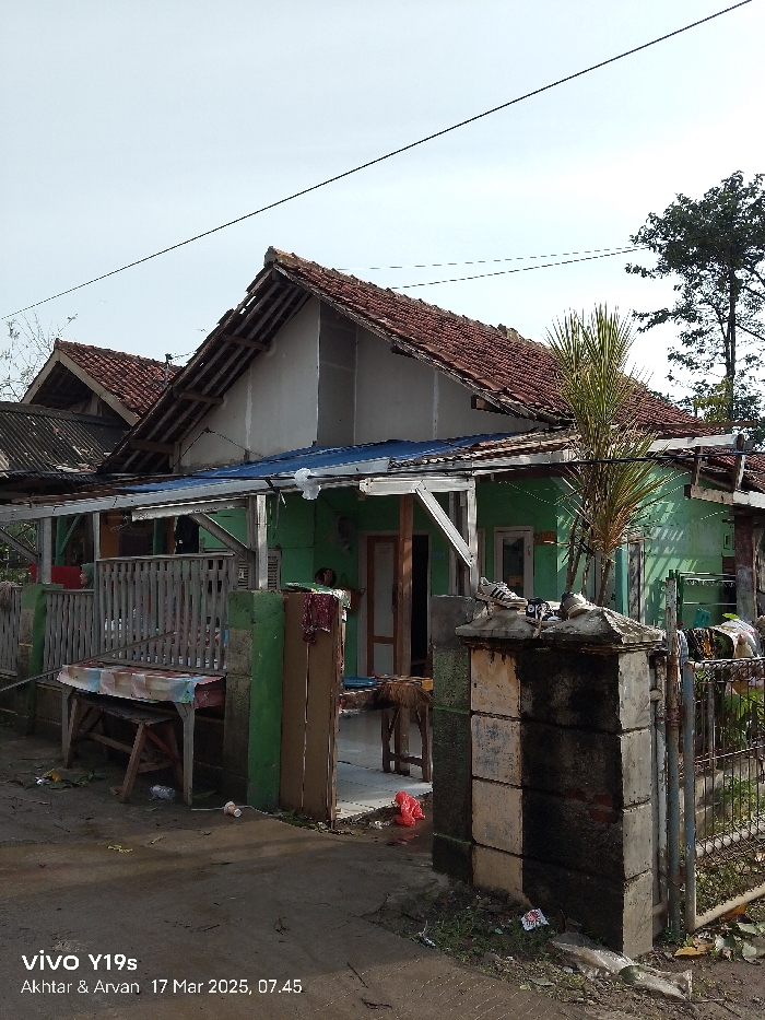 The image shows a small, green and white house with a wooden roof. The house is located in Anjatan, Indonesia, at coordinates -6.388 latitude and 107.93 longitude. There is a wooden fence in front of the house and a wooden table with a colorful tablecloth sitting in front of the fence. There is a palm tree growing next to the house. The ground is paved with concrete. The house appears to be in a state of disrepair, with some of the roof tiles missing. There are some items of clothing hanging on a clothesline in the background.