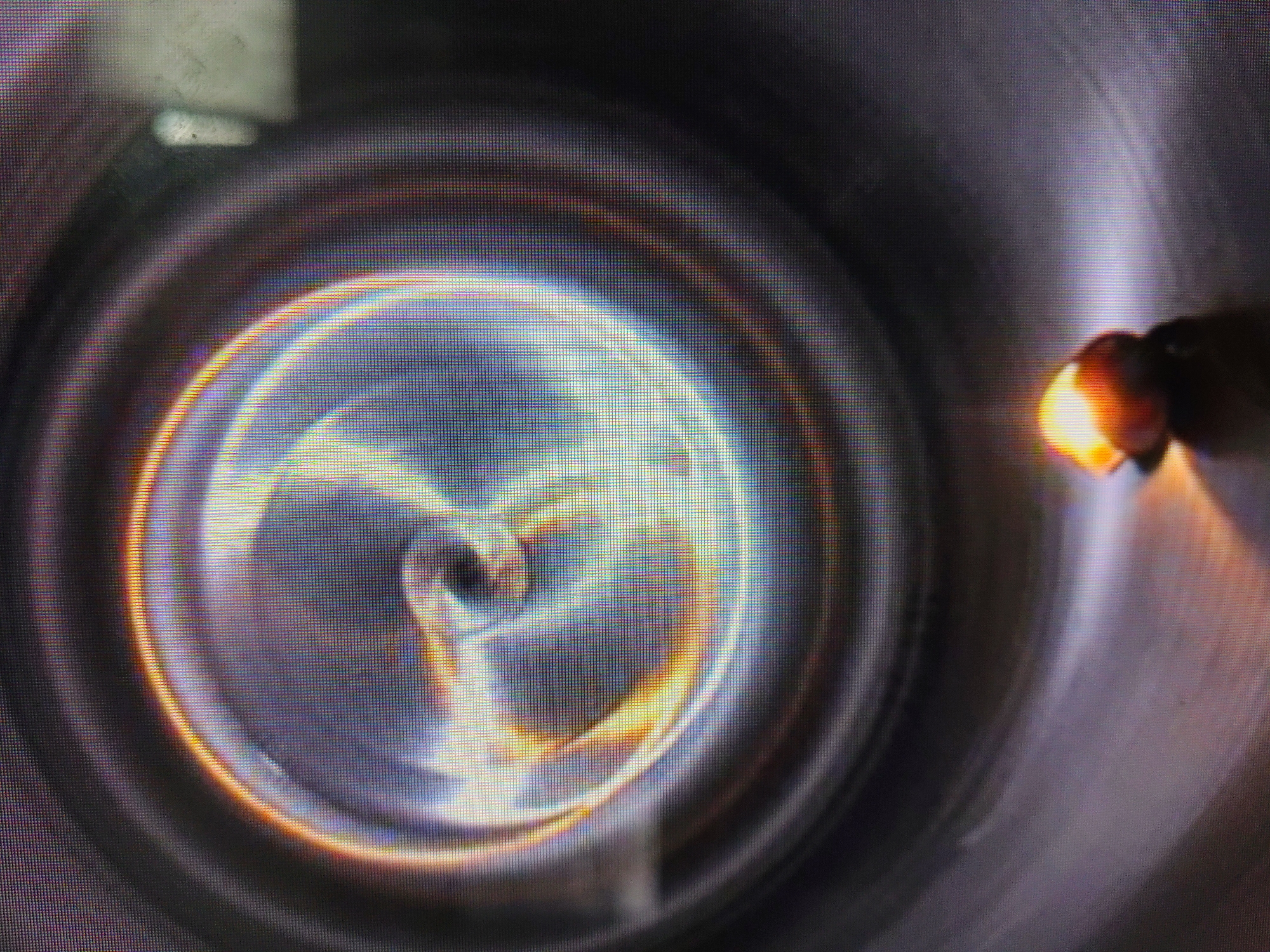 A close-up, pixilated image shows a light source emitting a swirling, rainbow-colored light pattern. The light pattern resembles concentric rings with dynamic streaks of yellow, white, and blue. In the foreground, a small, brightly lit, orange object is visible, casting a shadow. The background is dark and appears to be a reflective surface, possibly metal. The image is taken from a low angle, looking upwards into a circular opening or chamber. The lighting is intense and appears to be originating from within the chamber. The overall impression is one of scientific or industrial experimentation. The scene is located in Unknown, Unknown.