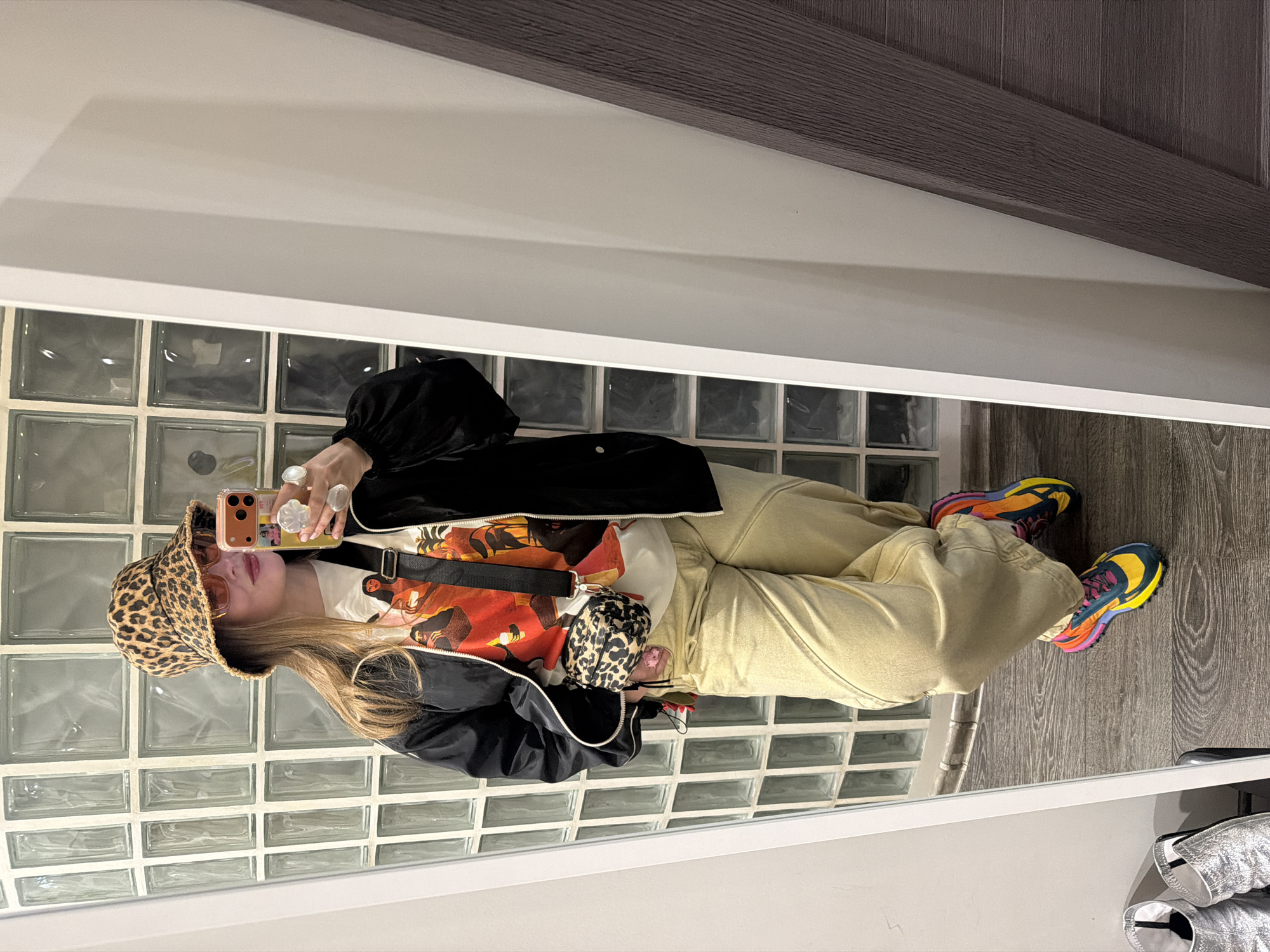 A person stands in front of a mirrored wall, holding a smartphone to take a picture of their reflection. The person is wearing a leopard-print bucket hat, sunglasses, a printed T-shirt under a black jacket, beige pants, and colorful sneakers. A patterned purse hangs across their body. The background includes a wall made of glass blocks and a wooden-paneled wall above. The floor is made of gray wood planks. The location is not specified, and there is no indication in the image that the location is in the city of Moscow, Russia.