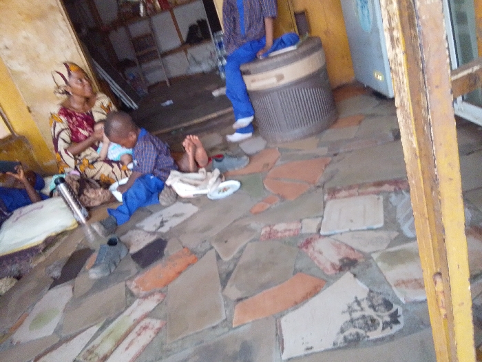 This image depicts an indoor scene in Hoche, Nigeria, likely within a commercial establishment or an open-fronted building. The floor is composed of irregularly shaped, mismatched concrete or tile pieces in shades of grey, orange, and white.
In the left foreground, a woman in a patterned dress and headwrap sits on the floor, holding an infant dressed in light blue. Beside her, a person lies reclined on a white mat, partially obscured, with a silver thermos flask nearby. In the center, a young boy in a blue plaid shirt and blue trousers sits on the floor, looking down at a white plate containing food. His bare feet rest on a white cloth, and a single dark shoe lies near him.
In the mid-ground, towards the right, another individual wearing blue trousers and a blue patterned shirt sits on a brown and beige ribbed plastic bin or cooler. Behind this person, a silver-grey refrigerator or freezer unit is visible against a yellowish-brown wall with peeling paint. Shelving with various items is present in the far left background. A prominent yellow wooden beam runs vertically in the right foreground, partially obscuring the view. The overall lighting suggests an indoor setting with ambient light.