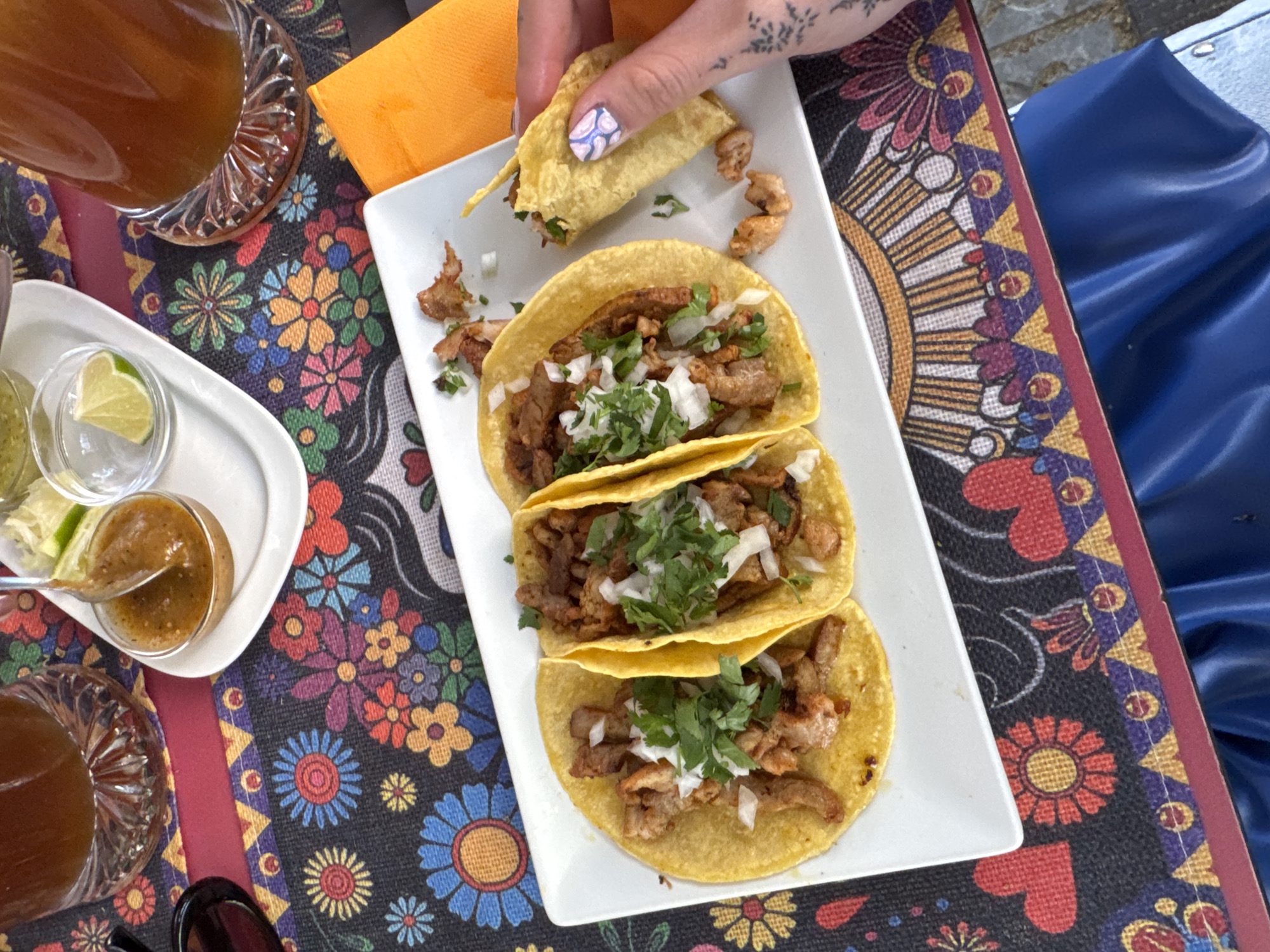 A slightly high-angle indoor shot shows a white rectangular plate with four small, yellow corn tortillas filled with meat, chopped onions, and chopped cilantro, with a hand holding one of the tacos. The plate sits on a colorful placemat with a floral and geometric pattern. To the left of the plate, there's a small white dish with two small bowls, one filled with salsa and the other with lime wedges. A spoon rests in the salsa. There are two tall glasses of liquid, with the edges of the glasses visible. An orange napkin is also visible near the top of the frame. The hand holding the taco has a tattoo and painted nails. To the right of the plate, there is a portion of a blue garment.