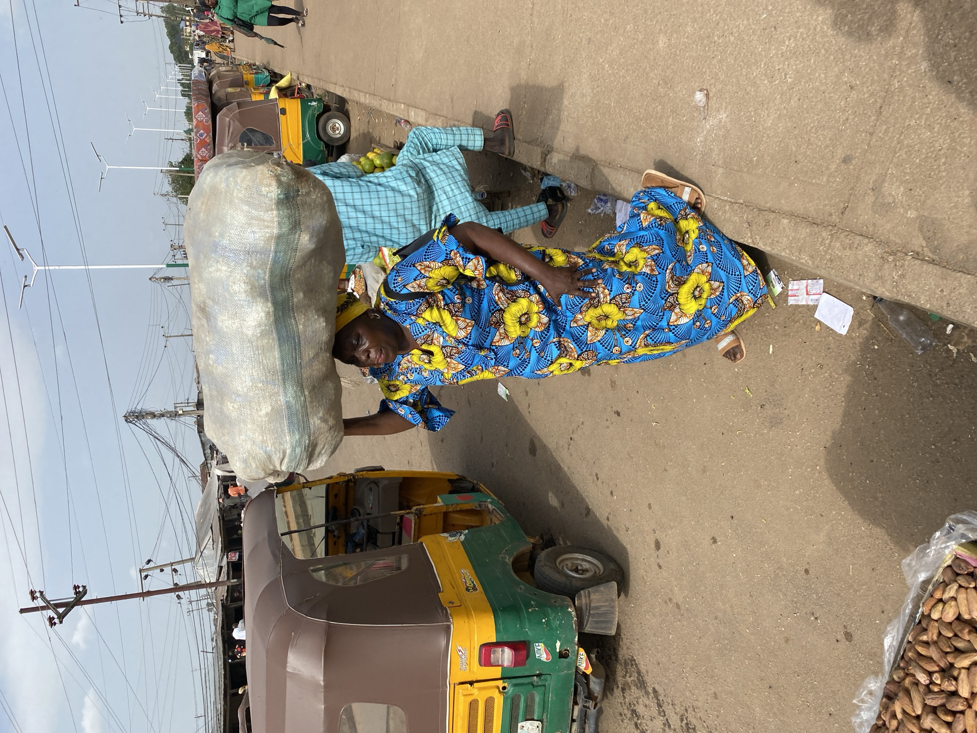 In this vertical, eye-level shot taken in Bukuru, Nigeria, two people are resting on the ground next to a busy street. A large, white burlap sack filled with goods rests on the shoulders of a person wearing a blue and yellow patterned dress. Their head is tilted back, and their eyes are closed, suggesting fatigue. To their left, another person wearing a blue checkered shirt and pants also lies on the ground, appearing to be asleep or resting.
The scene is set on a paved street with a concrete curb. An auto-rickshaw, a common mode of transport in Nigeria, is parked nearby. The auto-rickshaw is predominantly brown with green and yellow accents. The street is lined with various objects, including street lights, power lines, and what appear to be stalls or market structures in the background. Several other vehicles, including more auto-rickshaws, are visible further down the street.
Bags of produce, possibly groundnuts, are stacked in the lower right corner of the frame. The overall atmosphere suggests a hot, sunny day, as indicated by the bright lighting and the shadows cast on the ground. The people's relaxed postures and the presence of market-like elements point towards a market setting or a roadside resting area. The image captures a moment of rest amidst the hustle and bustle of daily life. There is no visible text in the image.
