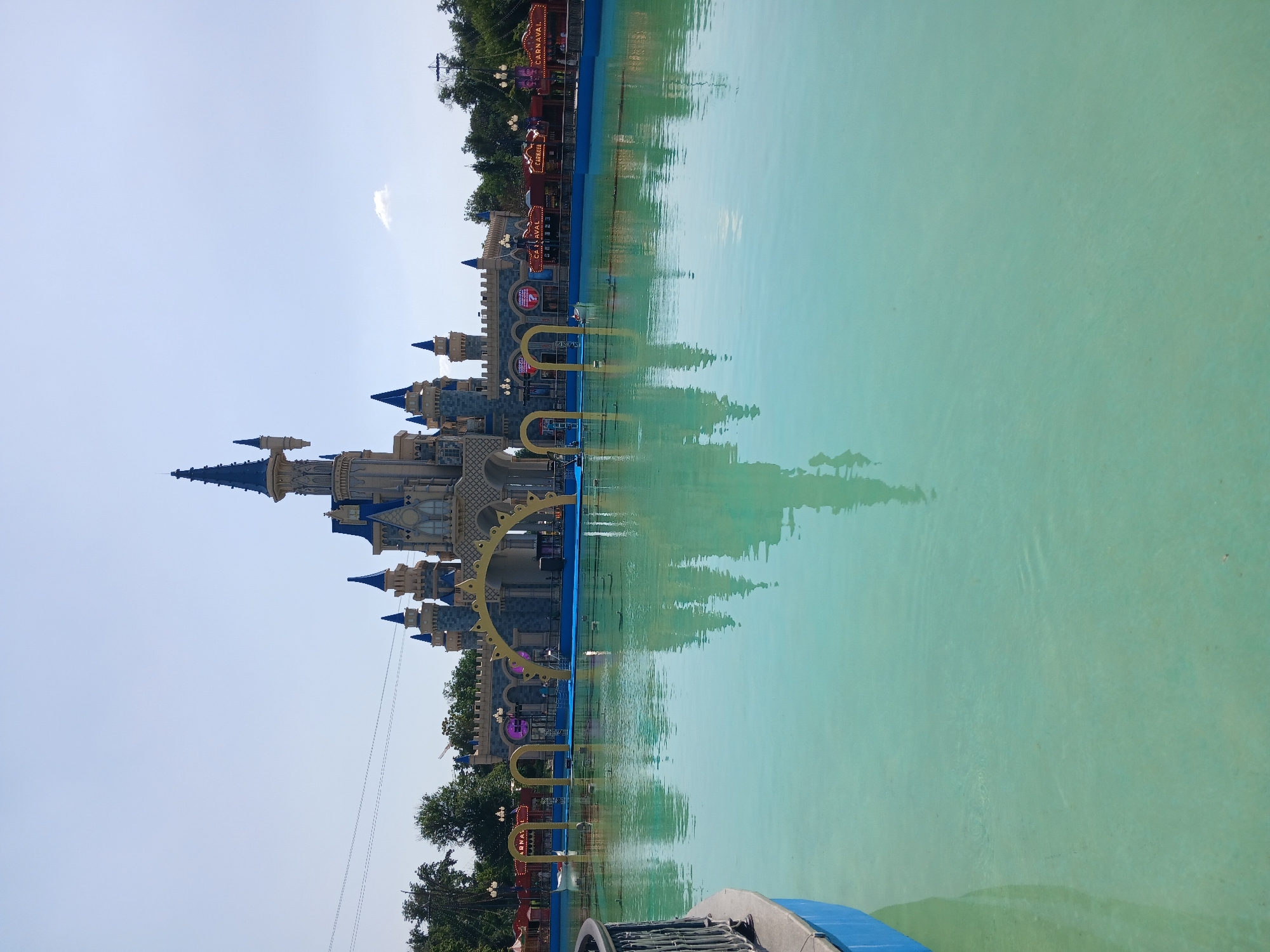 This image depicts a fairy-tale-style castle structure with blue turrets, situated next to a large, clear turquoise pool. The castle has multiple levels and architectural details, including arched windows and decorative elements. The water in the pool is calm, reflecting the castle and surrounding trees. On the far side of the pool, beyond the castle, there is a row of trees and some illuminated signs. One sign is clearly visible with the word "CARNAVAL" in red letters on a yellow background. The sky is a bright, clear blue, suggesting a sunny day. The overall scene evokes a sense of a theme park or amusement area, likely a water park given the prominent pool. The reflection in the water is a notable detail, adding depth and symmetry to the image. The lighting suggests it is daytime, possibly mid-morning or afternoon, due to the bright sunlight. The image's orientation is rotated 90 degrees counter-clockwise from a typical viewing perspective. Given the location context of Tashkent, Uzbekistan, this could be a recreational facility within the city. There are no people clearly visible in the image, focusing instead on the architectural elements and the serene water setting.