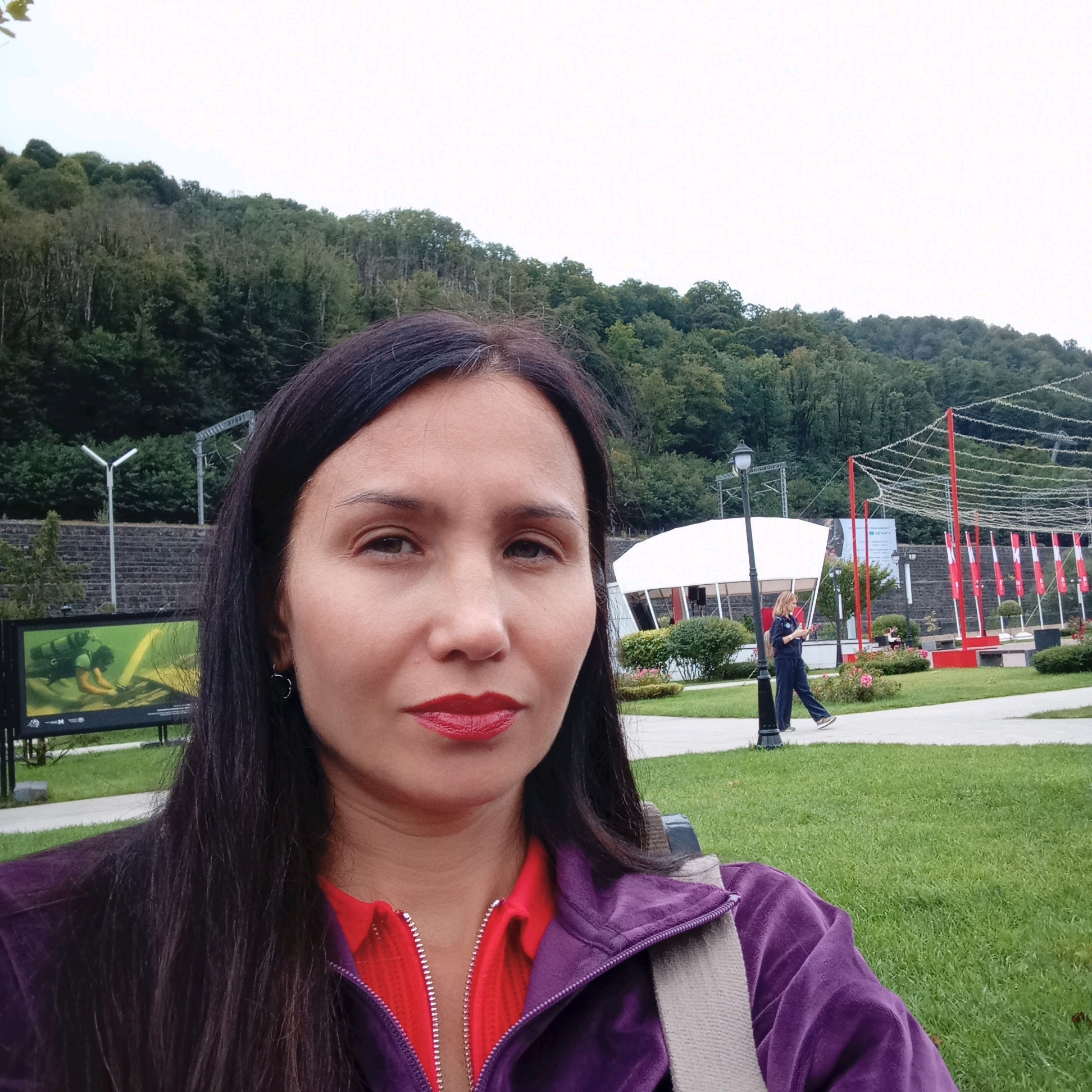 This is a medium shot, taken from a selfie perspective, of a woman in the foreground, with a park and a forested hillside in the background. The woman has long, dark brown hair and is wearing a purple zip-up jacket over a red collared shirt. She has on red lipstick and small hoop earrings. In the background, a white tent-like structure with angled glass panels is visible. To the right of the tent, a woman in blue athletic wear is walking on a paved path. Red banners hang from a structure to the right, and string lights are suspended overhead. In the far background, a densely forested hillside dominates the scene. The ground is covered in green grass, and there are some bushes and flowers. The location appears to be a park in Estosadok, Russia.