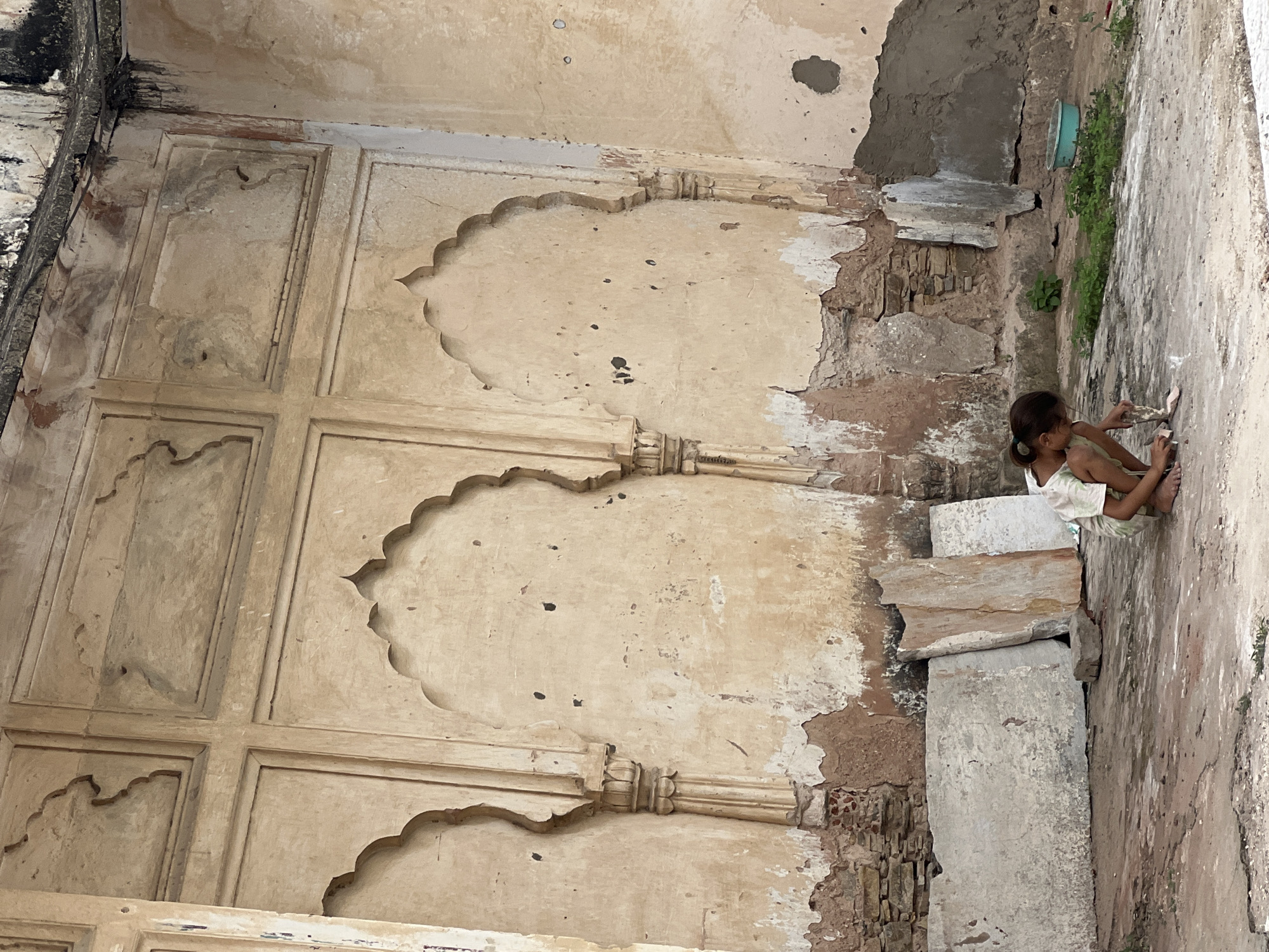 A young girl with dark hair pulled back in a ponytail kneels on the dusty ground in front of a dilapidated, ochre-colored wall in Pushkar, India. The wall features ornate arched niches and panels, some of which are peeling and showing exposed brickwork. Several large, flat stones lean against the wall behind the girl. She is wearing a light-colored t-shirt and shorts and is engaged in playing with small objects on the ground. The surrounding environment appears weathered and neglected, with patches of green vegetation growing amidst the debris.