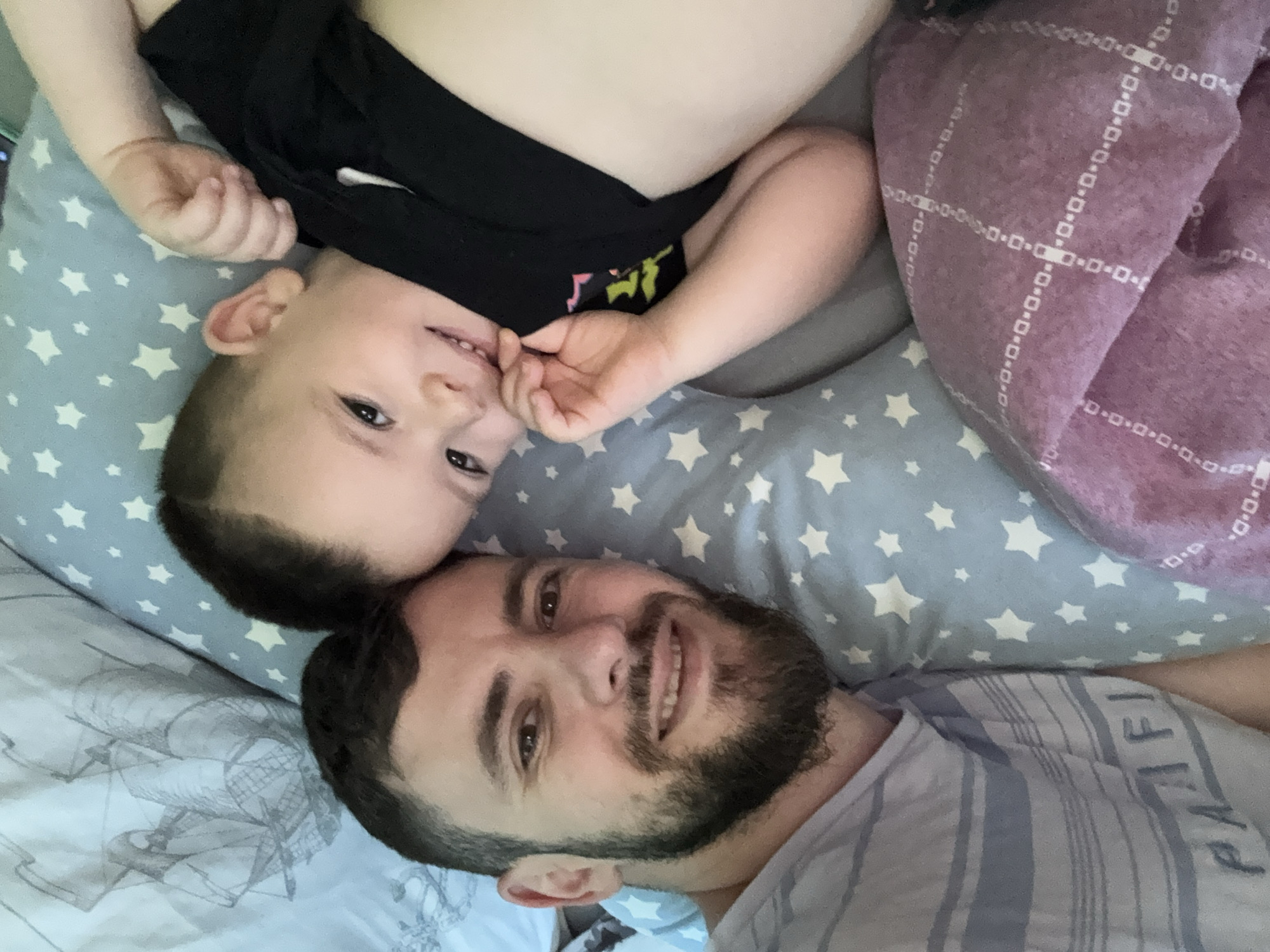 An adult male and a young child are depicted in a close-up selfie, positioned side-by-side, lying down on bedding. The scene is located in the city of Hadzhidimitrovo, Bulgaria. The adult male, positioned on the left foreground, has short dark hair, a dark full beard, and is smiling directly at the camera, revealing teeth. He wears a light-colored t-shirt with horizontal stripes, featuring the partially legible word "PACIFIC". The child, positioned to the right of the adult male and slightly further back, has short dark hair and is also smiling directly at the camera, revealing teeth. The child wears a black t-shirt with a partial light-colored graphic on the chest, and one hand is raised towards the mouth area. Bedding elements include a light grey sheet with a dark grey sailing ship pattern behind the adult male's head, a light blue pillow with white star patterns positioned between the two individuals, and a purple blanket with a white grid pattern of small squares and rectangles on the lower right. The lighting is soft and even across the faces of both subjects.