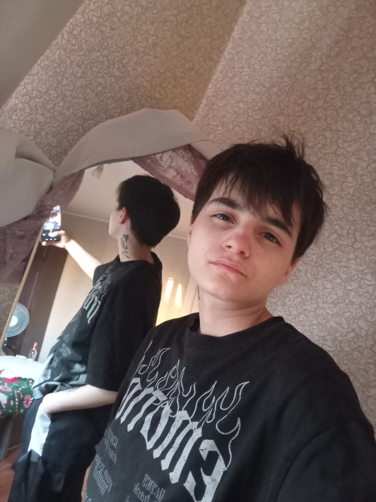 A person with short dark hair is taking a selfie in front of a mirror. The person is wearing a black t-shirt with a white graphic design. In the mirror, the person is visible taking the photo with a smartphone, also wearing a black t-shirt. A tattoo is visible on the neck of the person in the mirror. A partial view of a room includes wallpaper with a light-colored floral pattern. A fan, a table with a colorful tablecloth, and a bottle are reflected in the mirror. The location is possibly in a residential building in Naberezhnye Chelny, Russia.