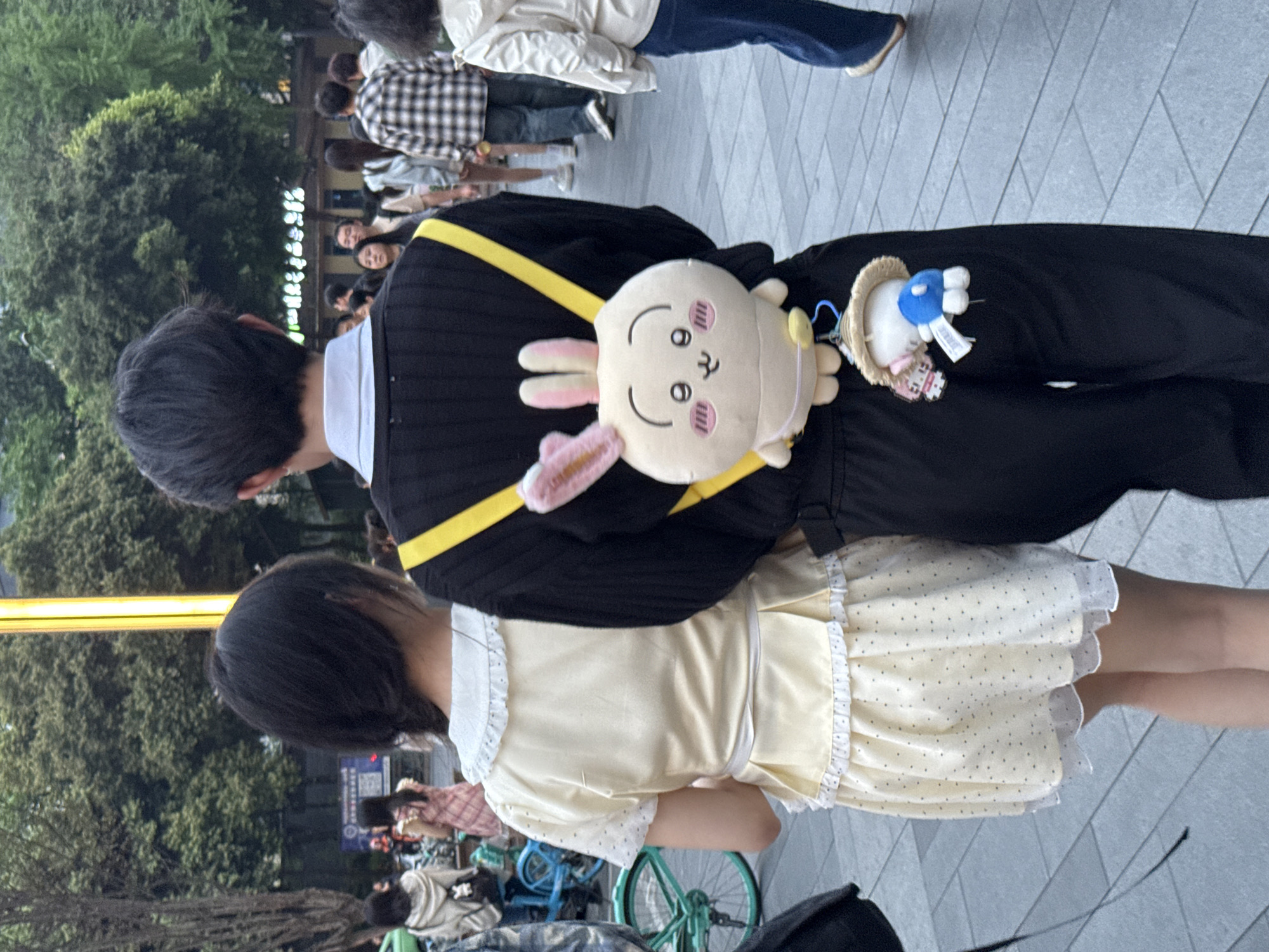 A medium shot depicts a street scene in the Jinjiang District of Chengdu, China, showing two people standing close to each other. A person with short, dark hair wears a black sweater with a yellow strap across the back and has a cartoon rabbit plush bag. The second person, also with short, dark hair, wears a light yellow dress with white frills. In the background, there is a crowd of people, trees, and a street with parked bicycles. The ground is covered in grey tiles.