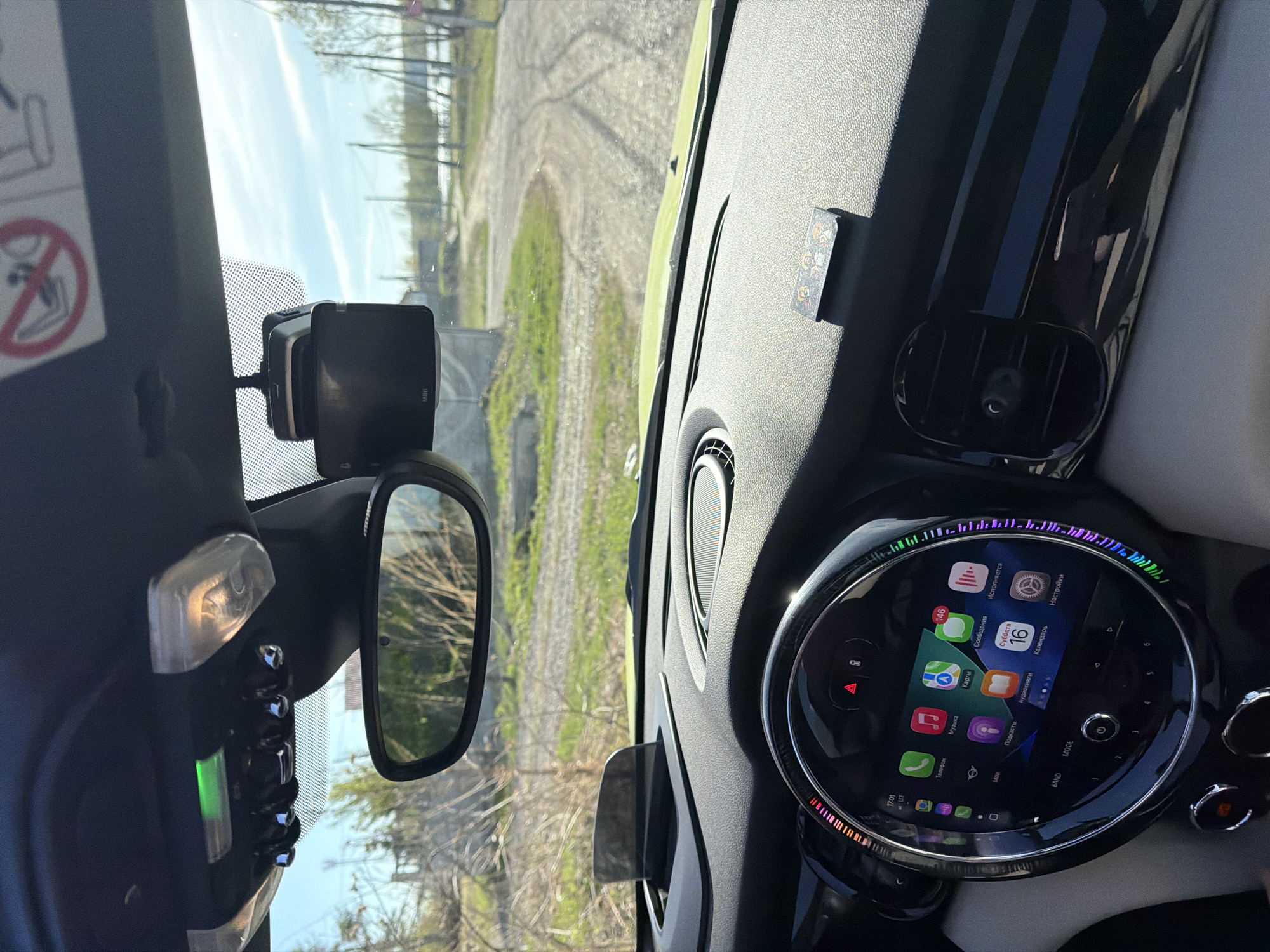 Here is a detailed description of the image:
**Location:** The image appears to be taken from inside a car, potentially in Zheleznogorsk, Russia (based on the OCR text with "LTE"). The location outside the car cannot be determined definitively.
**Scene:** The view is from the driver's seat, focusing on the car's interior and the road ahead. It's a clear day.
**Objects/Subjects:**
* Car Interior: Parts of the dashboard, steering wheel, rearview mirror, and center console are visible.
* Digital Display: A circular display screen shows various icons including those for apps such as Music, Maps, Messages, and Settings. The language on the screen appears to be Russian. Date and time is visible at the top of the screen.
* Road: The road outside appears to be gravel.
* Landscape: Trees and green areas flank the road.
**Actions:**
* The car is likely stationary or moving slowly, as the landscape visible through the windshield is somewhat clear.
* The digital display is active, indicating the car's systems are powered on.
**Interaction:**
* None visible, besides the use of the car's systems.
**Environment:**
* The environment outside the car suggests a rural or suburban setting, with a gravel road and natural vegetation.
**People:**
* No people are visible.
