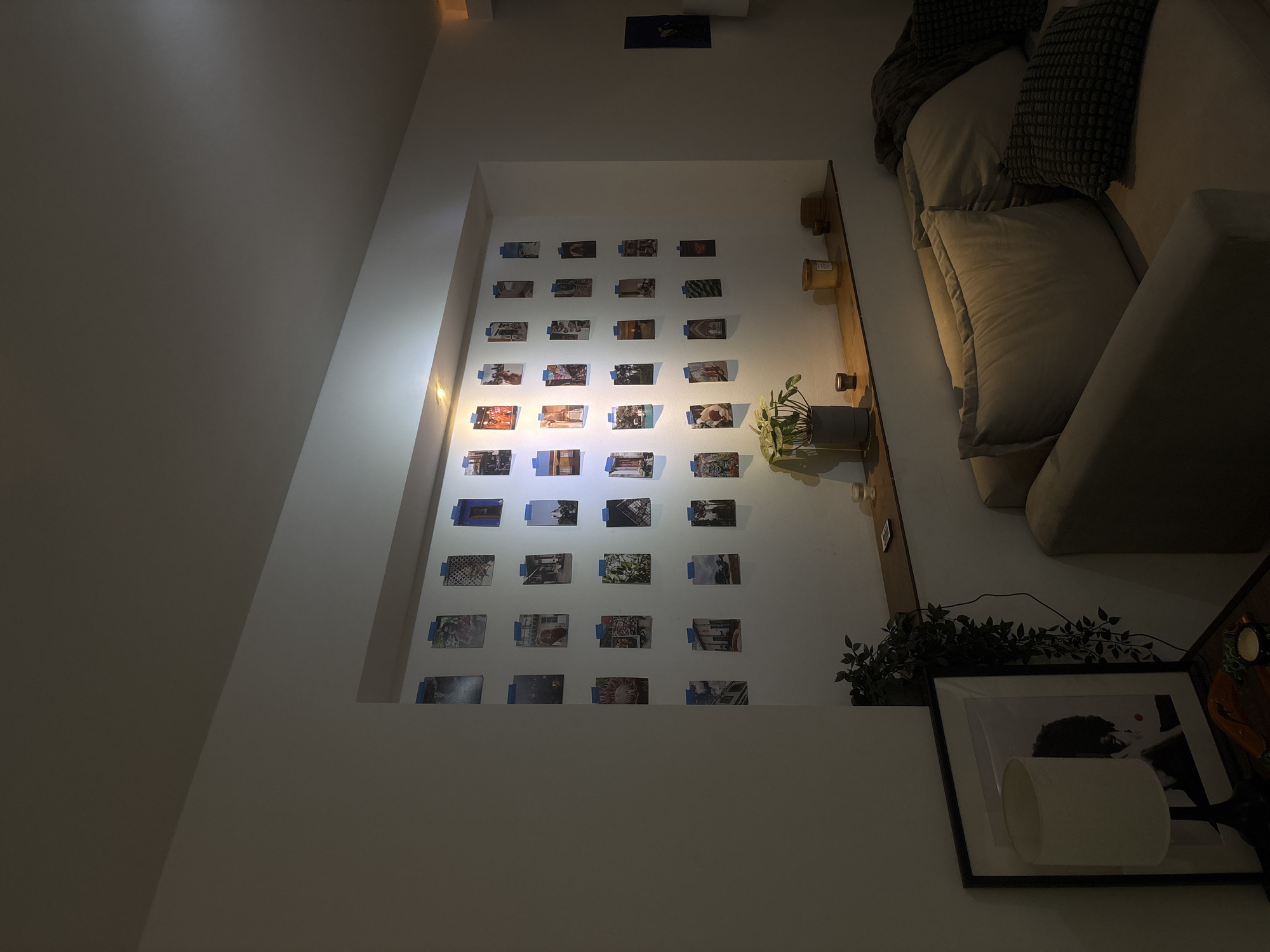 This is an interior shot of a living room. The walls are painted white, and a recessed wall feature displays 24 rectangular photographs attached with blue tape. A wooden shelf runs alongside the right edge of the recessed area, displaying decorative vases and a potted plant. A cream-colored sofa with throw pillows is placed next to the wall feature. A decorative plant and a framed black and white image are located next to the sofa, and a lamp is visible in front of the framed image.