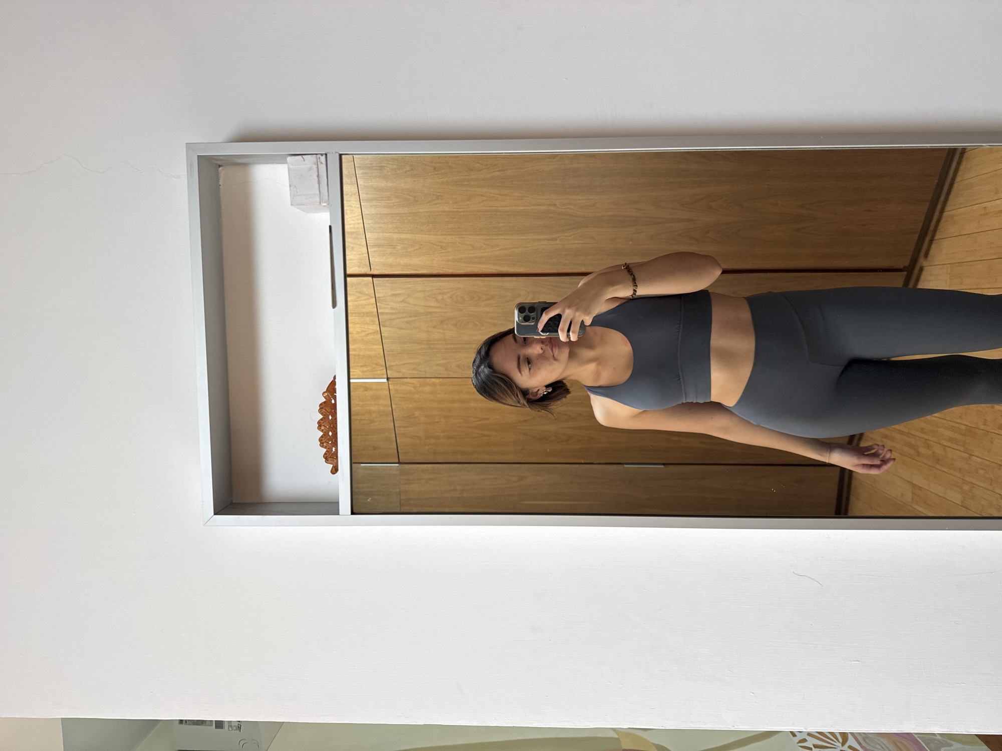 A young woman is taking a selfie in a mirror. She is wearing a gray athletic top and leggings. The mirror is framed in a silver-colored frame and is reflecting a wooden cabinet and a hardwood floor. The mirror is mounted on a white wall. A decorative object is in a shelf to the left of the mirror. The bottom part of a painting is visible in the bottom left corner. The location is San Bernardino Tlaxcalancingo, Mexico.