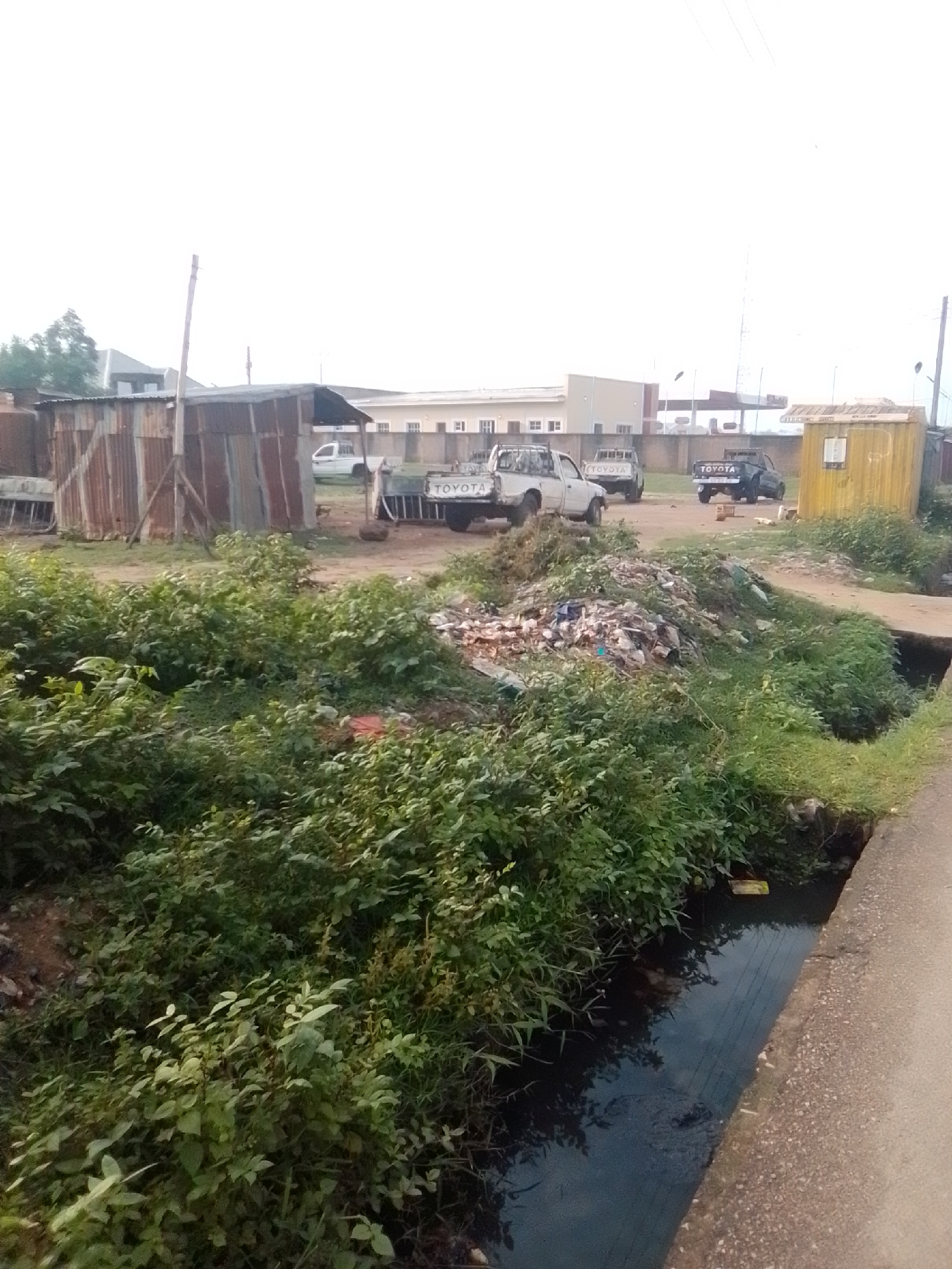 The image depicts an outdoor scene in the city of Gombe, Nigeria, characterized by a cluttered and somewhat dilapidated environment. In the foreground, a substantial pile of garbage, composed of plastic bags, paper, and other refuse, is visible, adjacent to a narrow, dark, and murky water channel running along a paved road. Dense green foliage encroaches on the edges of the garbage pile and the water channel.
In the middle ground, several pickup trucks are parked on a dirt surface. Two are clearly identifiable as Toyota models, one white and one dark-colored. Behind the trucks, a collection of corrugated metal structures, appearing to be makeshift shelters or workshops, stand. One of these structures is a rusty, lean-to style shed supported by wooden poles. Further in the background, a modern building with multiple windows and a fence is visible, suggesting a more developed area adjacent to the current scene. A yellow shipping container, likely used as a small shop or office, is positioned to the right of the trucks. The sky is overcast and bright. The overall impression is one of an area experiencing challenges with waste management and infrastructure.
