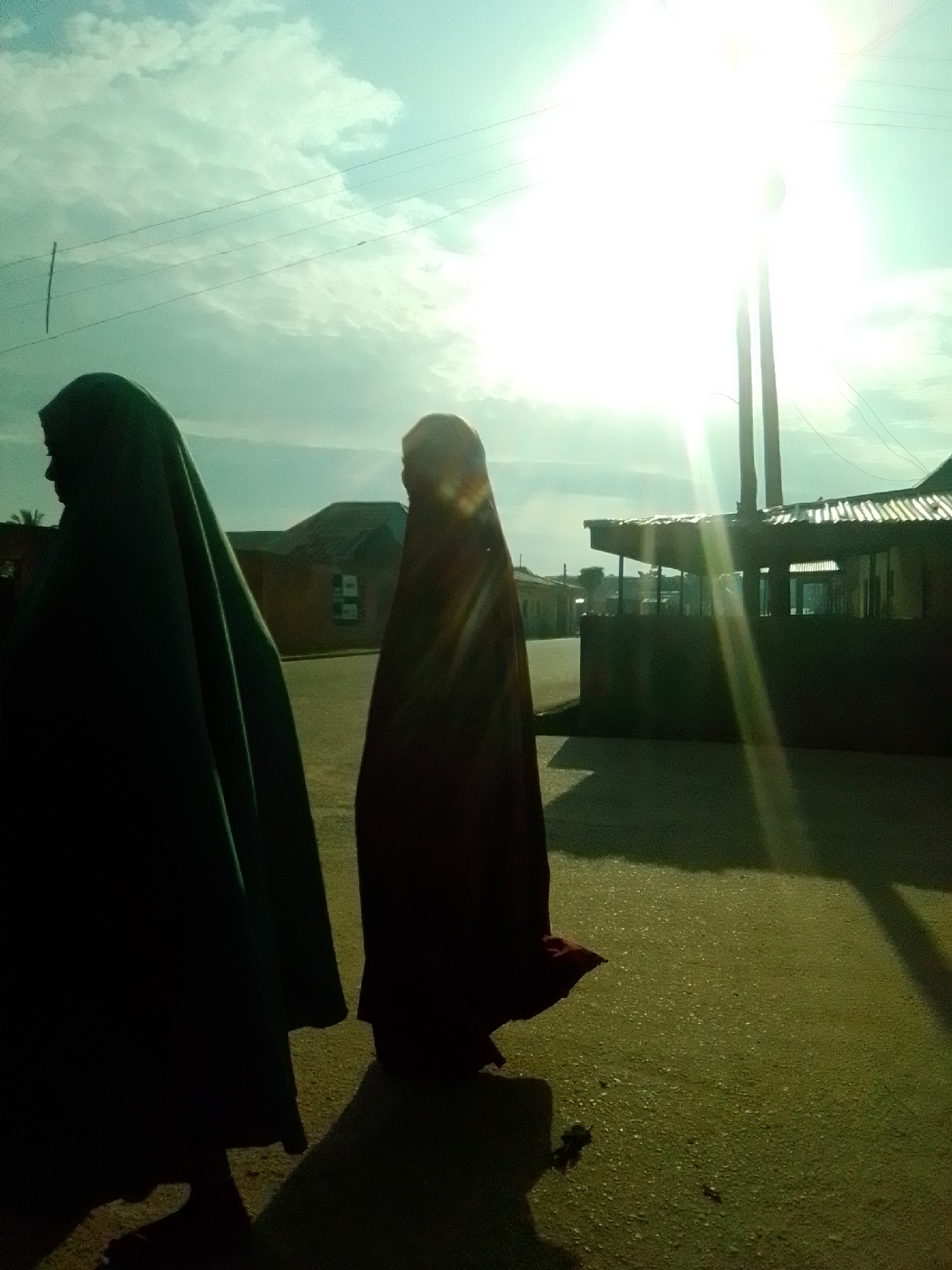 Two individuals are walking on a paved surface, possibly a road or sidewalk, in Liji Hill, Nigeria. The person on the left is wearing a dark green garment that covers their head and body, with only a profile of their face visible. The person on the right is also cloaked in dark fabric, obscuring their features. They are walking away from the camera, towards buildings in the background that appear to be houses or shops. The sky is bright with a strong sun casting lens flares and a light haze. Power lines are visible strung across the sky. The overall impression is a bright, somewhat hazy day with strong sunlight.