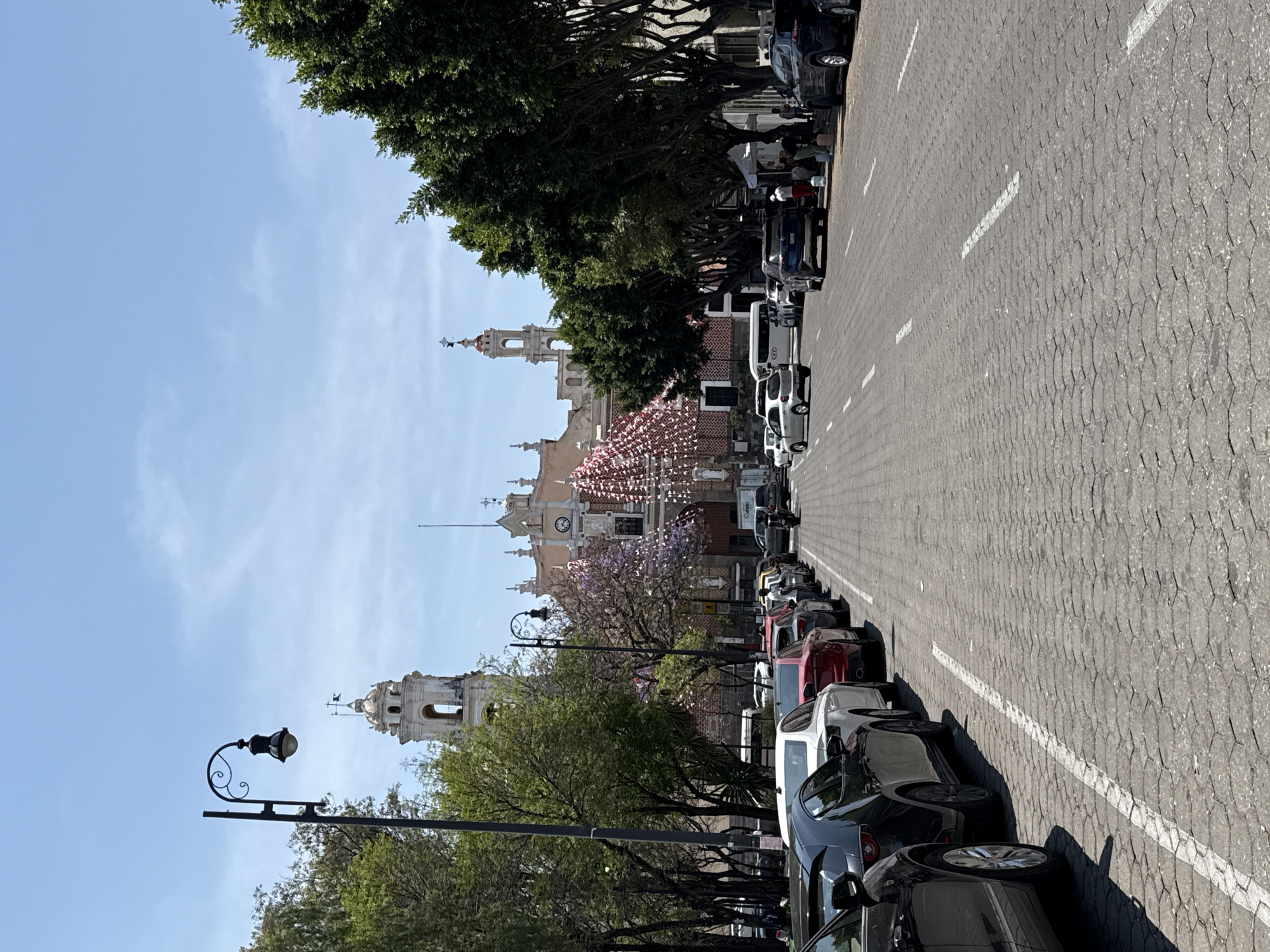 This full-shot street scene, captured on a sunny day, appears to be set in a historical or colonial city, consistent with Puebla, Mexico. The image is oriented vertically (rotated 90 degrees clockwise).
In the background, a grand, light-colored building with ornate architecture dominates the scene. It features multiple bell towers, domes, and a prominent clock face, characteristic of a large church or cathedral. Pinkish or reddish decorative banners, possibly papel picado, are strung across the area in front of this building, suggesting a festive atmosphere or celebration.
The foreground and midground feature a wide street with a textured, possibly cobblestone or patterned concrete, surface. A row of vehicles, including sedans, SUVs, and a white van, are parked bumper-to-bumper along the right side of the street. Strong shadows cast by the vehicles indicate bright sunlight, suggesting it is midday or early afternoon.
On the left side of the street, a tree-lined sidewalk is visible. The trees have lush green foliage, and one prominent tree in the midground displays purple blossoms, likely a jacaranda. A decorative black streetlight with an ornate bracket stands in the left foreground. A few indistinct figures of people can be seen near the parked cars and on the sidewalk, but no specific activities are clearly discernible. The sky is clear and blue with minimal, wispy clouds. No visible text is clearly legible in the image.