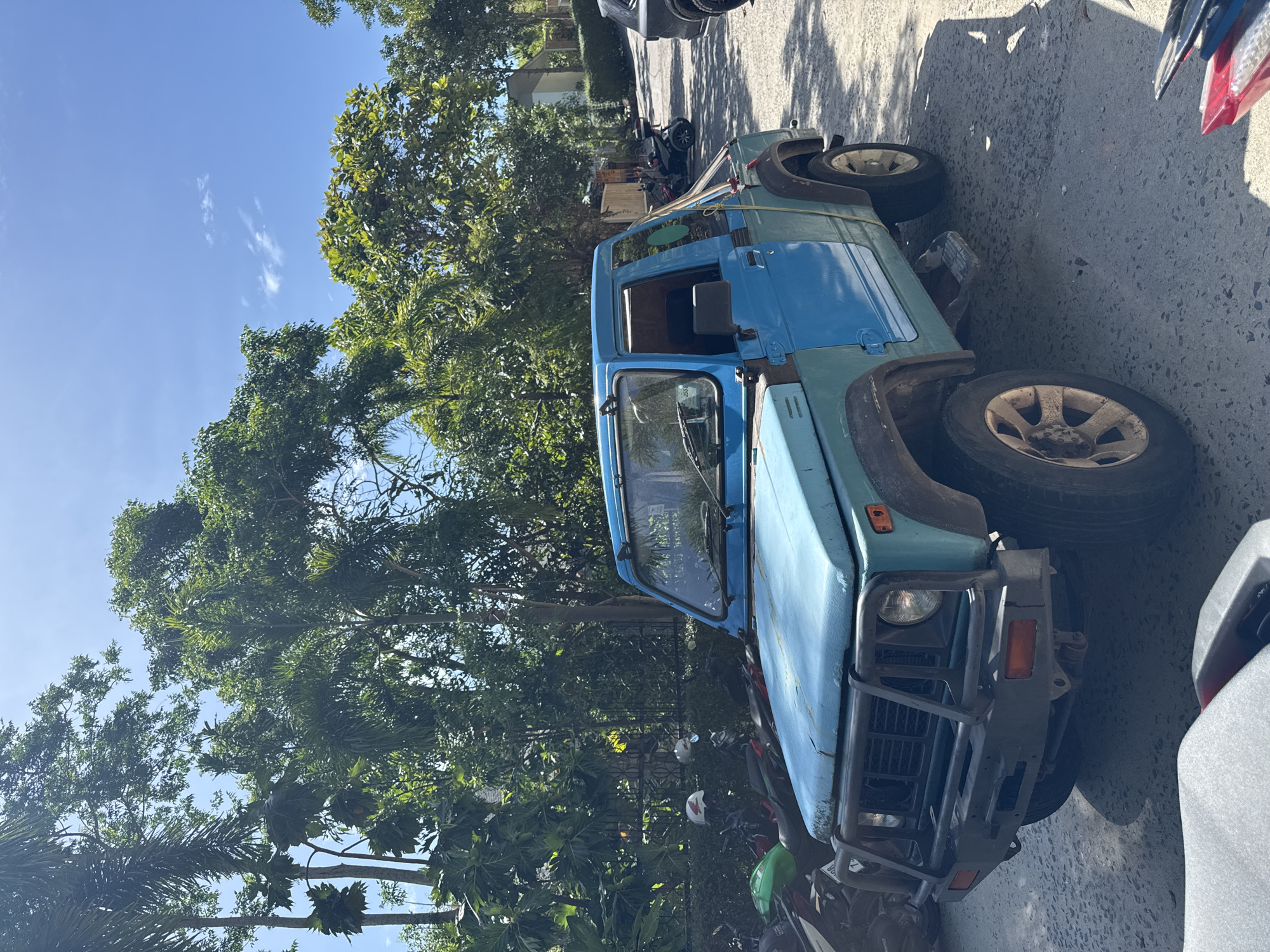 In ตำบลเกาะพะงัน, Thailand, a light blue, older model Suzuki Samurai is parked on an asphalt road. The vehicle has a black bull bar on the front and a step plate below the driver's side door. Behind the Samurai, several motorcycles are parked, some with helmets on them. Tall trees with lush green foliage surround the area, casting shadows on the ground. The sky above is clear blue with a few wispy clouds. In the background, a building with a light-colored facade is visible among the trees. The scene appears to be a sunny day in a tropical environment.