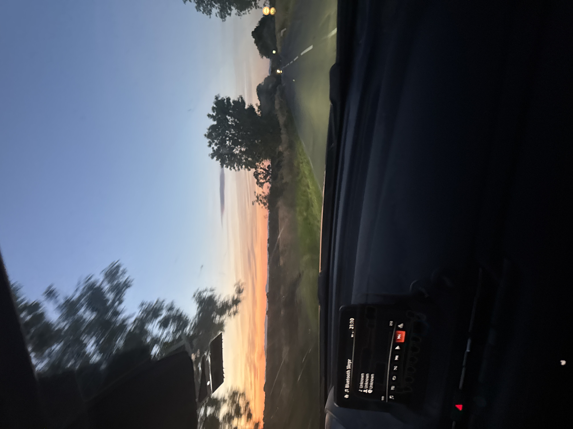 The image depicts a sunset viewed from inside a moving vehicle on a road near Elhovo, Bulgaria. The sky transitions from blue to orange hues, and the sun is setting over a landscape with trees and fields. The road ahead is illuminated by the vehicle's headlights, and a dividing line is visible. A dashboard screen shows music playing via Bluetooth. The interior of the vehicle is dark, with only portions of the dashboard and steering wheel visible.