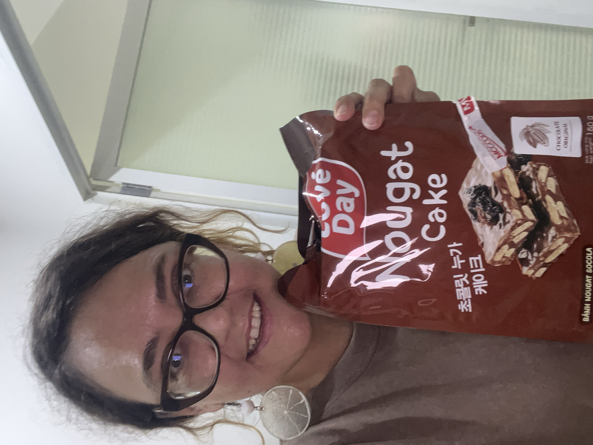 A woman with curly dark hair, wearing glasses and large circular earrings, smiles at the camera while holding a package of "Love Day Nougat Cake." The package is dark brown with red and white lettering. The woman is wearing a brown shirt. The background appears to be an indoor setting with a pale green sliding door. The scene is likely in Nha Trang Ward, Vietnam, given the text on the package which includes "BÁNH NOUGAT SOCOLA" and Vietnamese characters.