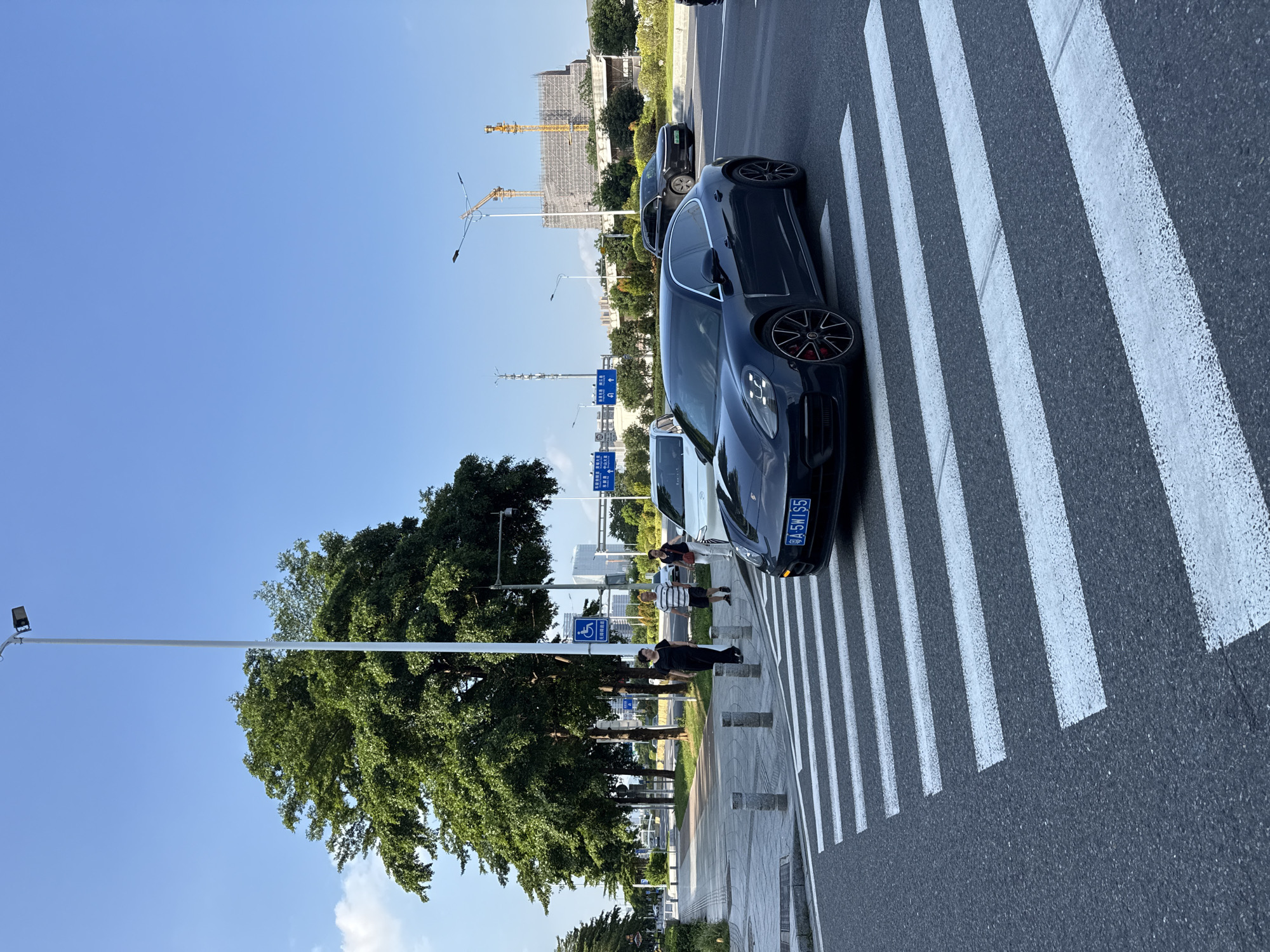 A dark blue Porsche Panamera drives across a marked pedestrian crossing on a sunny day in Haizhu District, China. The car, with license plate "A 5W1S5", has red brake calipers visible. To the left of the car, a white sedan is stopped at the crossing. Several pedestrians, two women and two men, are walking on the sidewalk to the left of the crossing. A blue sign with a wheelchair symbol is posted on a pole. Mature trees with lush green foliage line the sidewalk, and a tall white lamppost is visible. In the background, a multi-story building under construction with yellow cranes is present. The sky is clear and blue. The road surface is asphalt with white painted stripes for the pedestrian crossing.