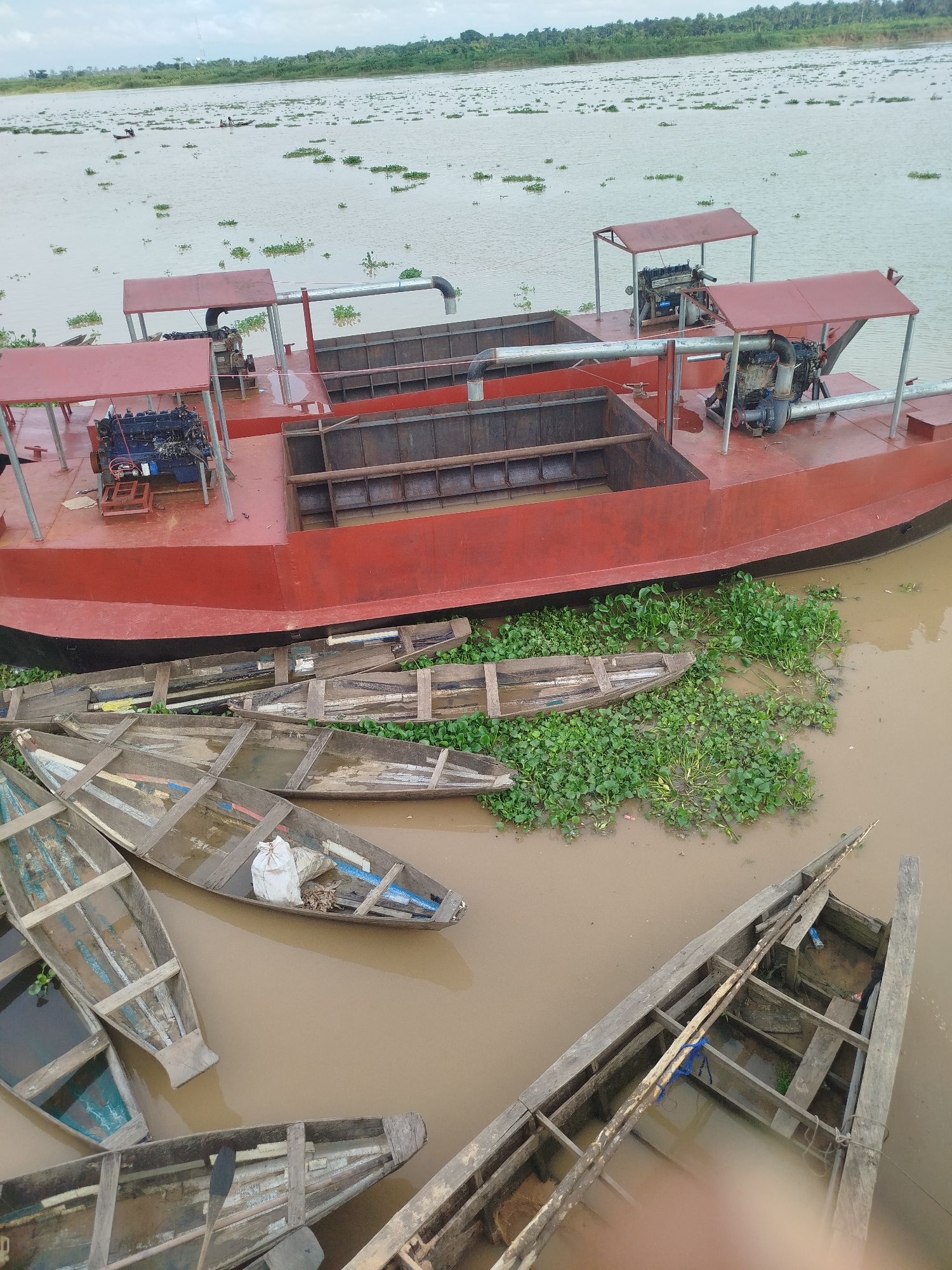 This media file shows a large red barge docked in a muddy river, likely in Yenegoa, Nigeria. The barge has two prominent engines with red roofs over them and large pipes extending from the back. Several smaller wooden canoes are moored alongside the barge, some filled with water, others with debris. One canoe has a white bag inside. Dense green aquatic plants are floating on the surface of the river, interspersed with the canoes and around the barge. In the distance, the river stretches out, dotted with more aquatic vegetation. The horizon shows a line of lush green trees, suggesting a tropical environment. The sky is overcast, indicating potentially cloudy or rainy weather. The scene appears to be a working dock or riverbank, with the barge likely used for transport or material handling. There are no visible people in the immediate foreground or on the barge, though two small figures can be seen in distant canoes on the river. The overall impression is one of a functional, perhaps somewhat weathered, riverine environment.