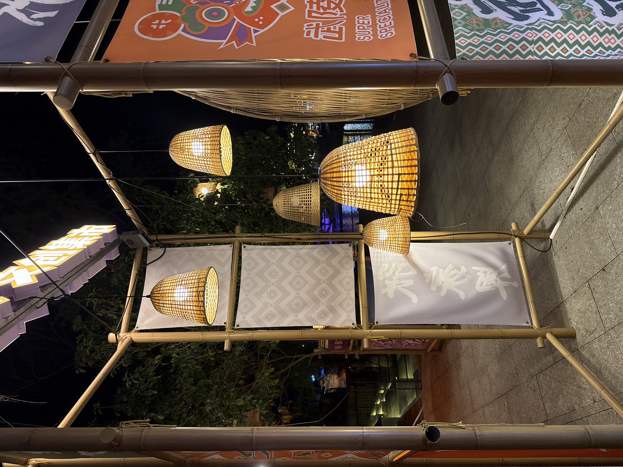 The image captures a vibrant outdoor setting at night, characterized by a meticulously constructed bamboo framework adorned with various decorative elements. Numerous bamboo poles crisscross, forming an overhead structure from which woven, bell-shaped lanterns emit a warm, inviting glow. These lanterns, made from interlaced natural fibers, are the primary light source, illuminating the immediate area and casting gentle light on the surrounding foliage.
Several banners or flags are suspended from the bamboo frame. One prominent orange banner displays English text "SUPER SPECIALTY" alongside Chinese characters. The characters "武陵" (Wǔlíng) are clearly visible, and the overall text likely spells "武陵源" (Wǔlíngyuán), directly referencing the location context in China. Other banners are white or light grey, featuring different designs: one has a repeating geometric pattern in light grey, another displays bold, white Chinese brushstroke characters that appear to read "喫快活" (chī kuàihuó), meaning "eat happily," and another shows "喫蝦蟹" (chī xiā xiè), translating to "eat shrimp and crab." These texts suggest the setting is a food market or dining area.
The scene is entirely devoid of people, allowing the focus to remain on the festive decorations and the intricate bamboo craftsmanship. The time of day is clearly night, with the dark sky providing a stark contrast to the illuminated elements. Lush green trees and bushes are visible in the background, adding natural texture to the artificial structures. The ground beneath appears to be paved with stone or concrete tiles, visible in sections. The overall mood conveyed is one of traditional festivity and a welcoming atmosphere, likely for an evening market or gathering in Wulingyuan, China.