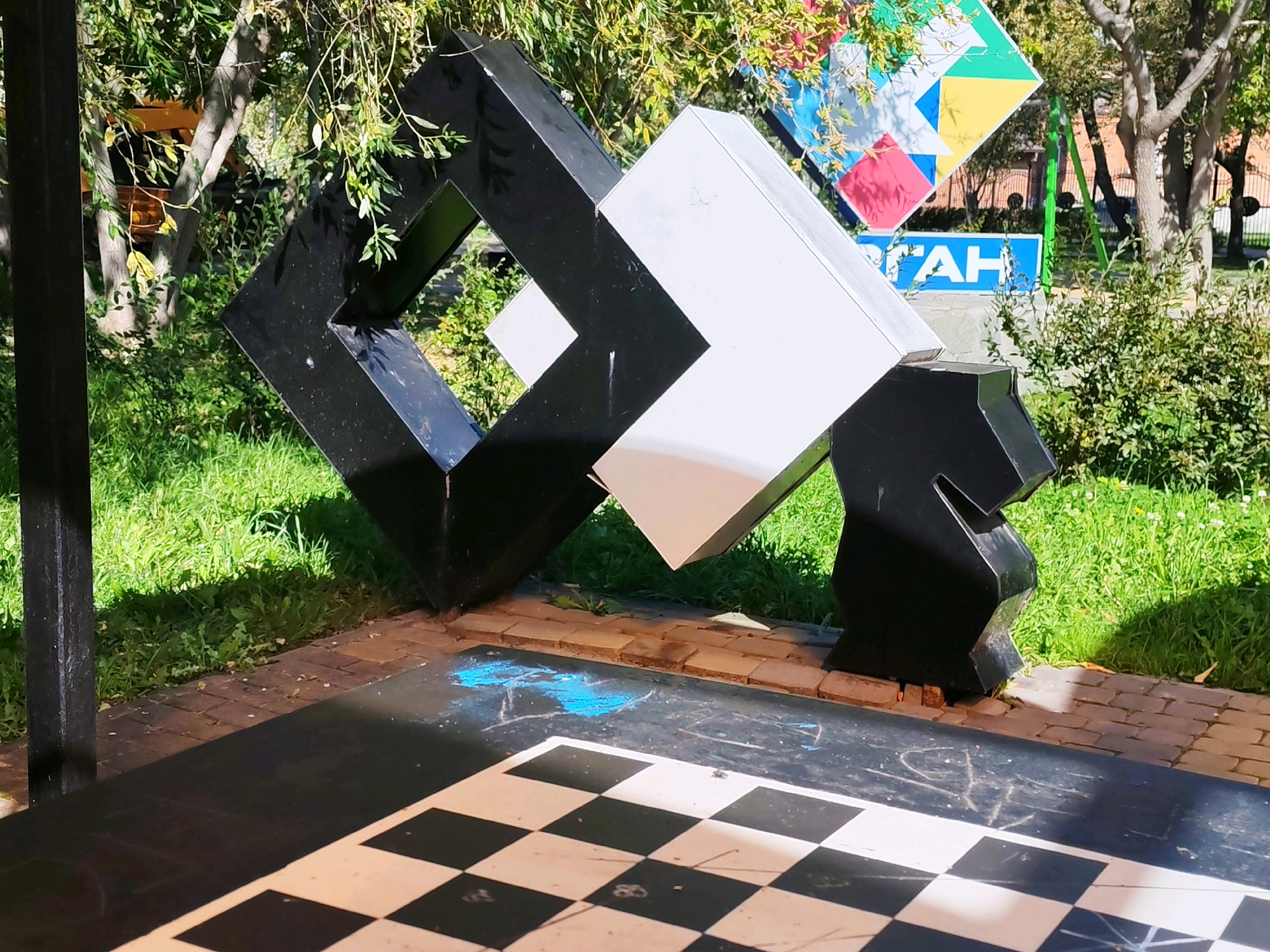 An outdoor, daytime, eye-level shot features a large, abstract sculpture composed of interlocking black and white geometric shapes, resembling stylized chess pieces. The sculpture is positioned on a paved area with a checkerboard pattern in the foreground, suggesting a connection to chess. Behind the sculpture, a patch of green grass and trees with dappled sunlight create a natural backdrop. In the background, a colorful mosaic-like sign with geometric shapes and text is visible, along with a blue sign with white Cyrillic lettering. The overall scene is bright and appears to be set in a public park or recreational area.
